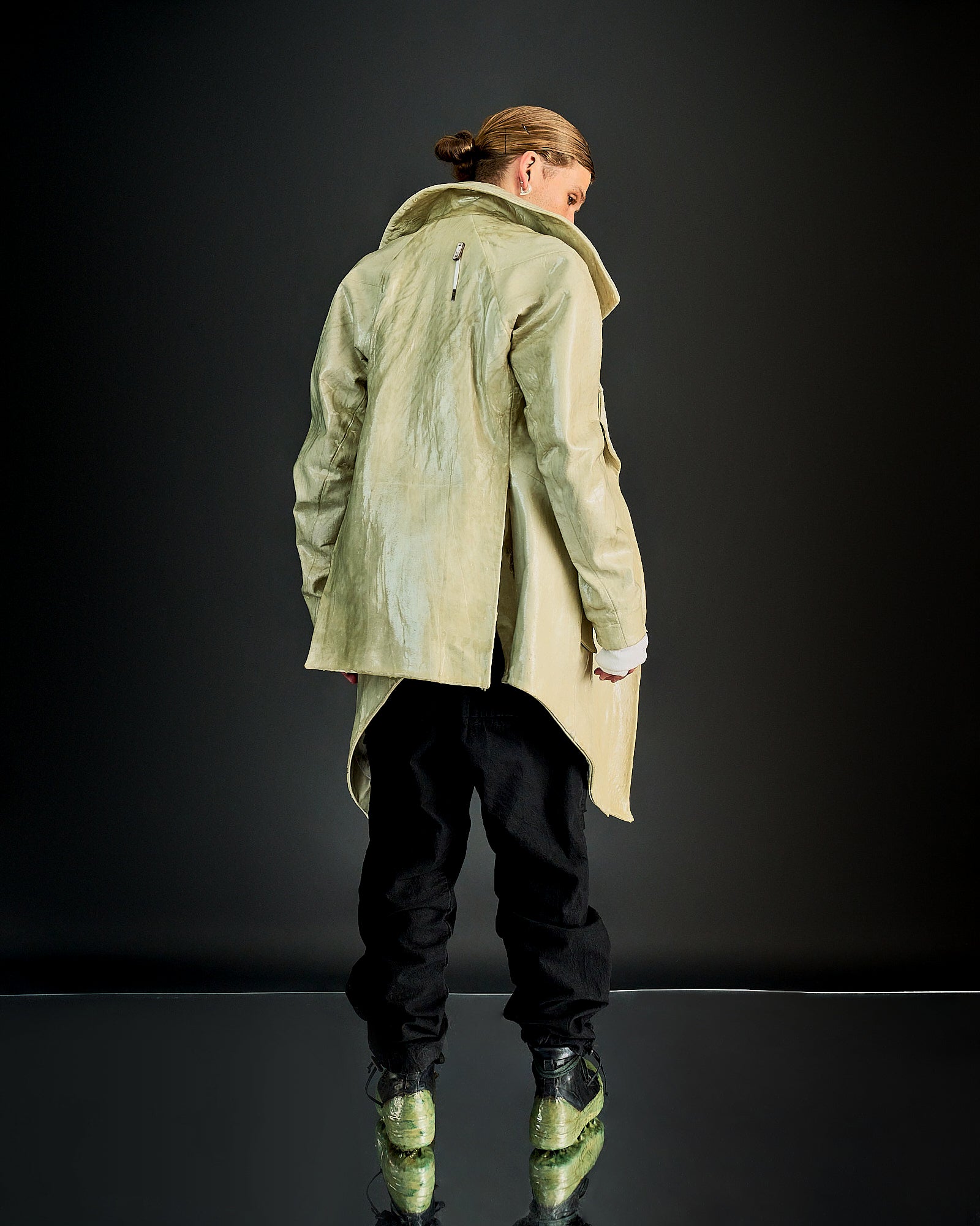 Person in a light Boris Bidjan Saberi coat faces away, wearing black pants and green transparent-sole shoes. The setting is a reflective, dark-toned surface.