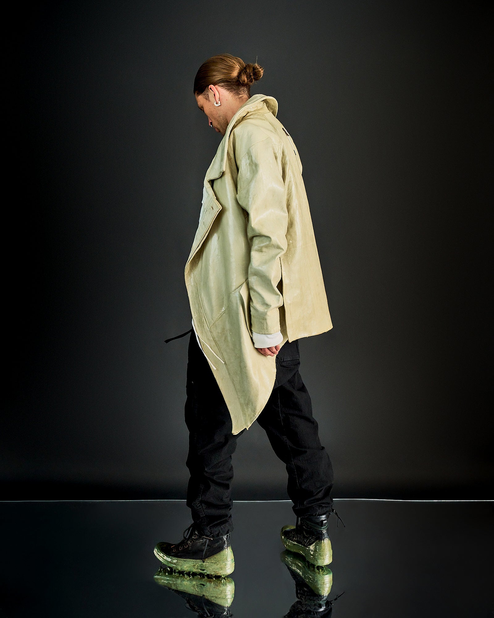 A person with a bun hairstyle wears a beige Boris Bidjan Saberi trench coat and black pants, standing on a reflective surface with neon green-soled shoes against a dark background.