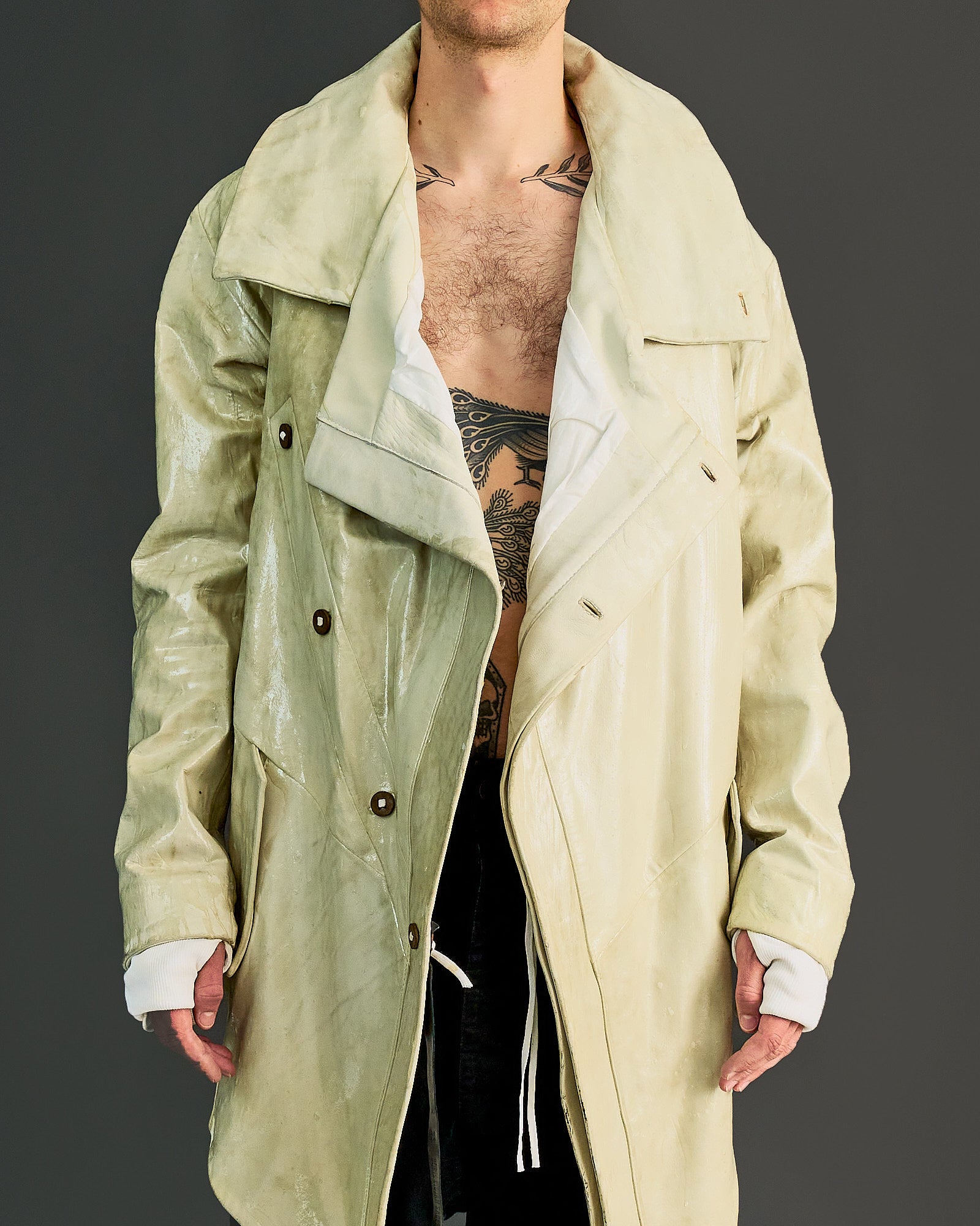 A person wearing an oversized, glossy beige Boris Bidjan Saberi trench coat with a partially open front, revealing a tattooed chest and torso. 