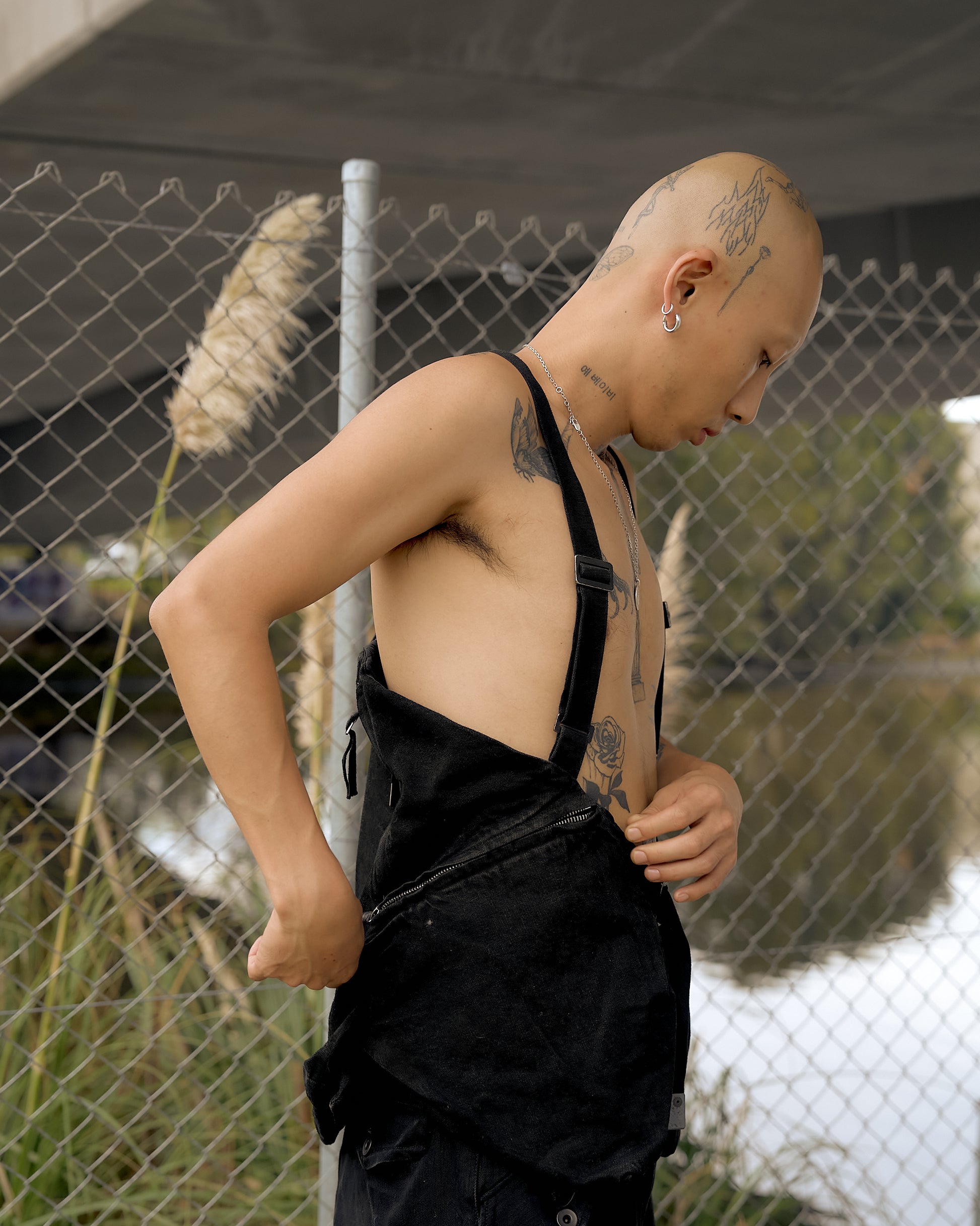 A bald person with tattoos, wearing black Boris Bidjan Saberi gilet stands by a chain-link fence near a river. 