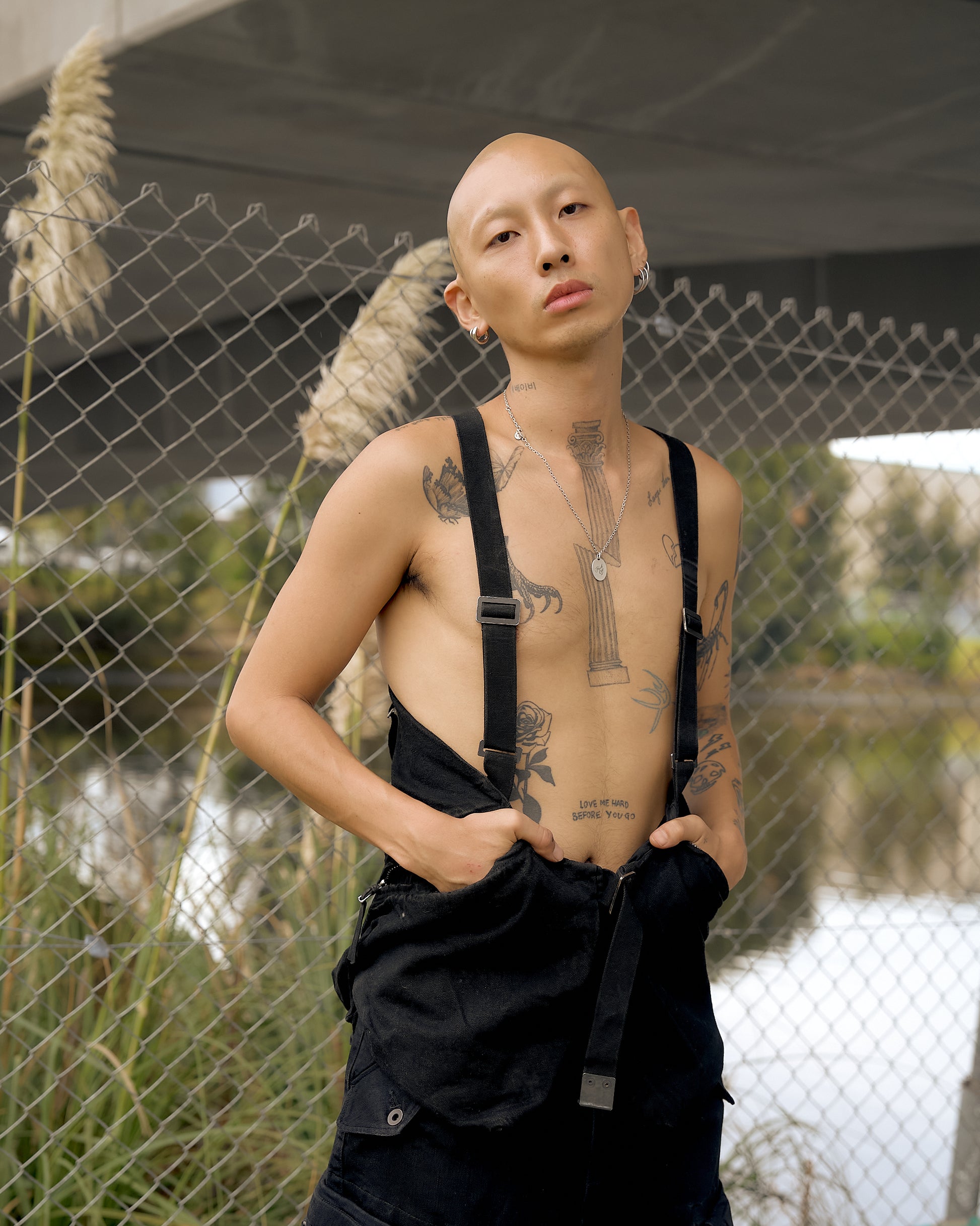 A person wearing a sleeveless waxed cotton Boris Bidjan Saberi gilet with adjustable belt straps and multiple pockets, standing near a wire fence with plants in the background.