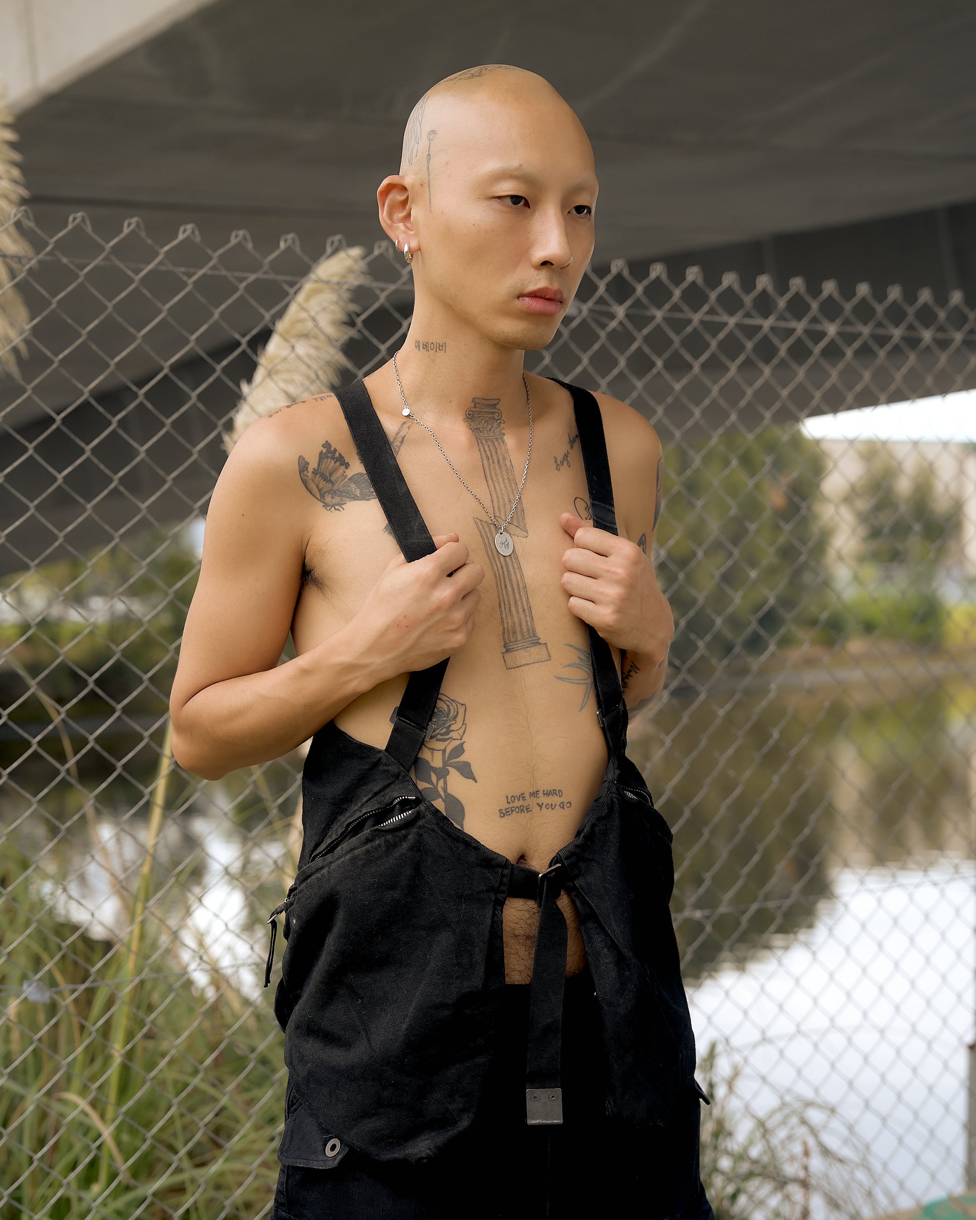 A tattooed, shirtless person stands near a chain-link fence, wearing a Boris Bidjan Saberi gilet. 