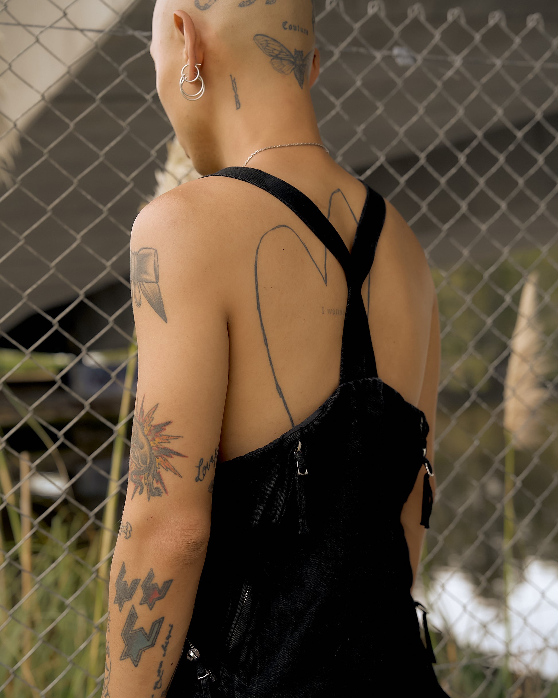 A person with tattoos, including a butterfly and star, stands by a chain-link fence. They wear a black Boris Bidjan Saberi gilet