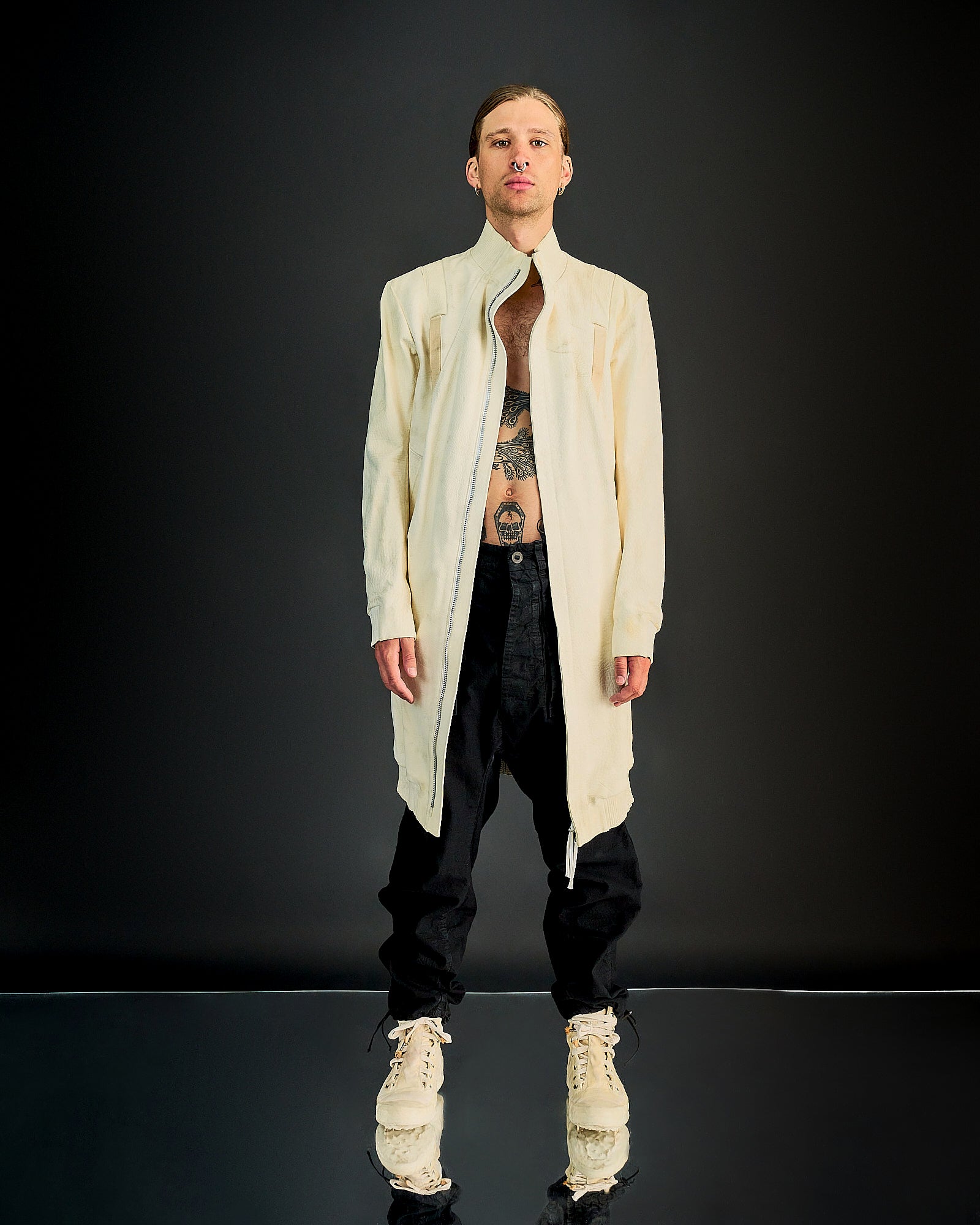 A person wearing a beige, knee-length, zip-up Boris Bidjan Saberi jacket made of Yak leather with a structured fit and ribbed cuffs. The jacket features a front zip and side pockets, and is accented with silver-aglet lace on the back.
