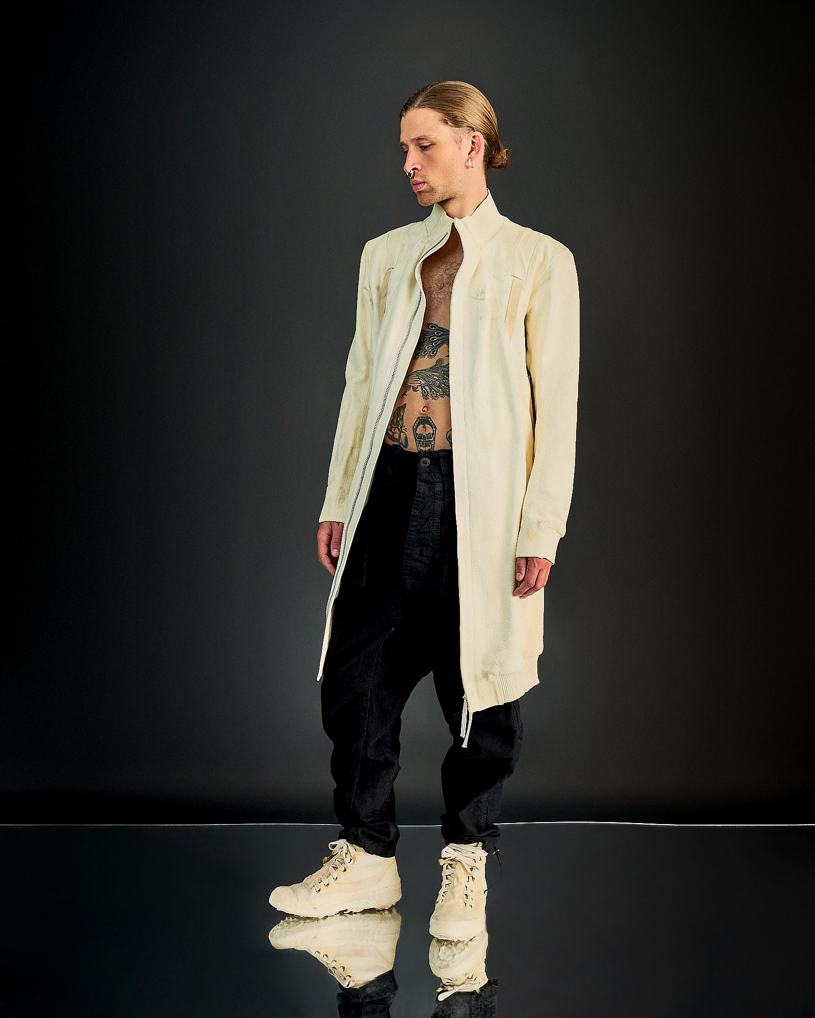 Person in open cream-coloured Boris Bidjan Saberi coat revealing torso tattoos, paired with black pants and matching high-top sneakers.