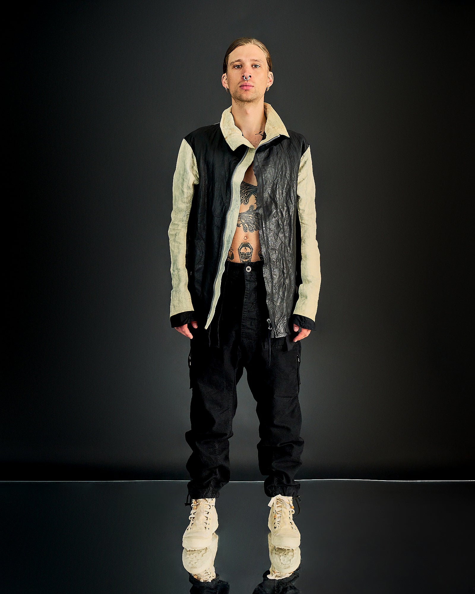 Person stands against dark backdrop, wearing a black and beige leather Boris Bidjan Saberi jacket paired with black pants and cream boots. 