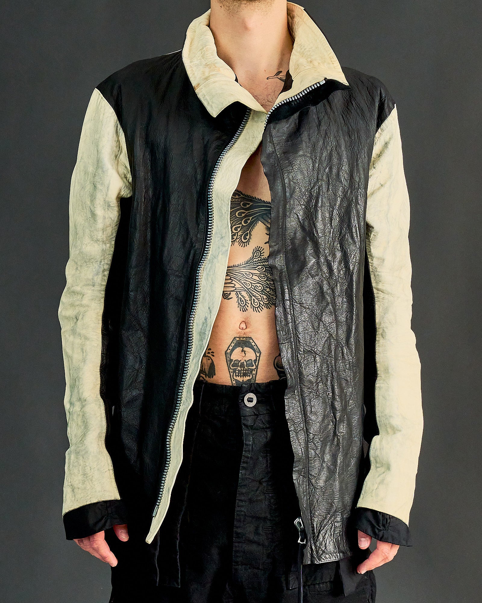 A person wearing a two-toned, open leather Boris Bidjan Saberi jacket with a visible tattooed torso.