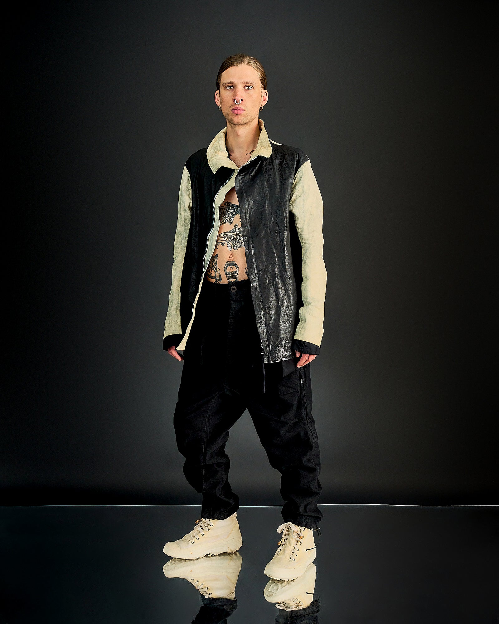 A person stands on a reflective surface wearing a black and beige Boris Bidjan Saberi jacket, black pants, and white boots. 