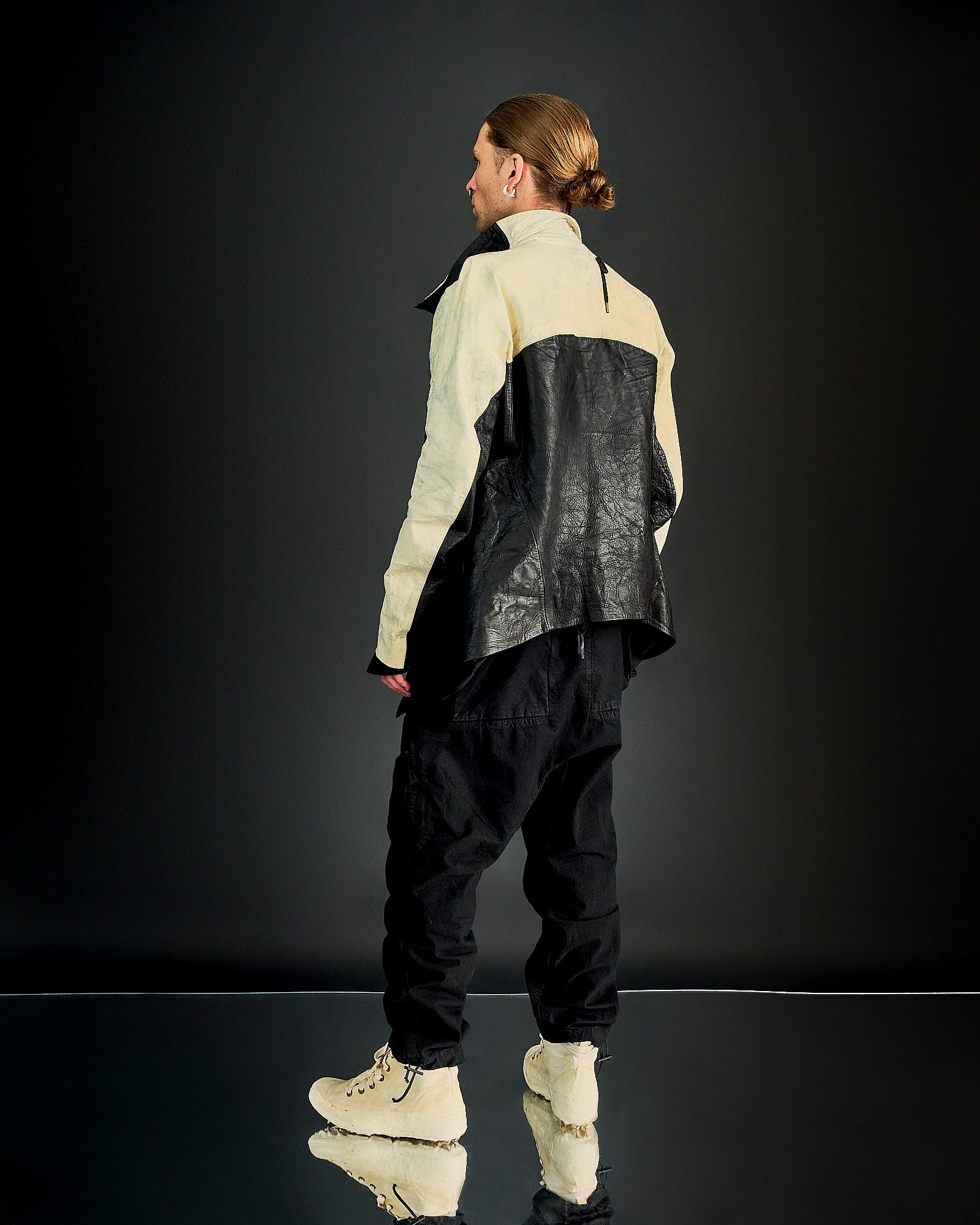 A person with long hair in a bun, wearing a two-tone Boris Bidjan Saberi outfit with a black leather back and white front, baggy black pants, and beige boots.