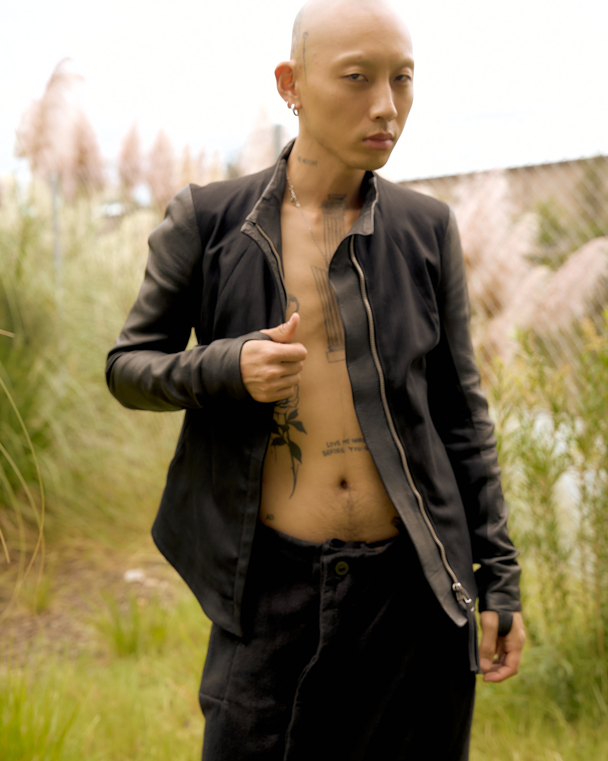 A bald person stands confidently in a grassy field, wearing a black Boris Bidjan Saberi leather jacket open to reveal chest tattoos. 