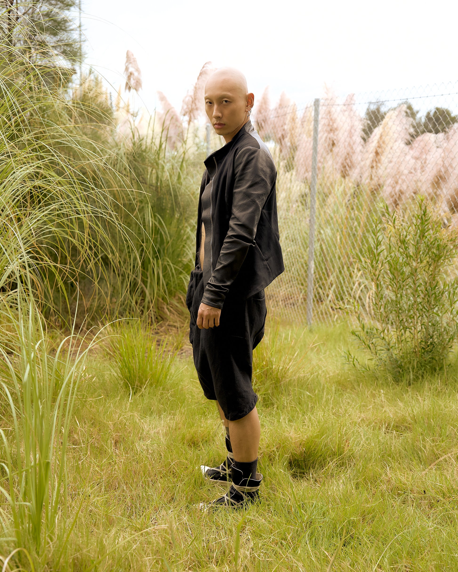 A person with a shaved head stands confidently in a grassy field, wearing a black Boris Bidjan Saberi  outfit and sneakers. 