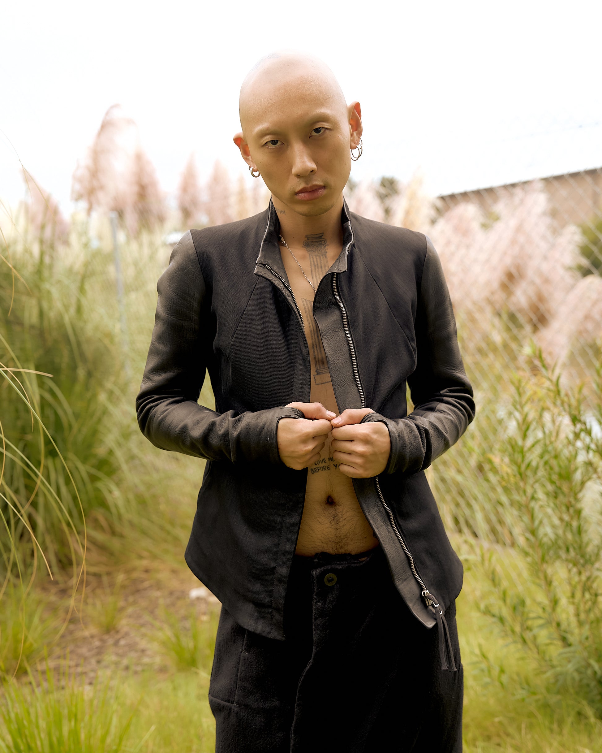 A person wearing a black dual panelled Boris Bidjan Saberi leather jacket with a round neckline and long sleeves, standing outdoors with tall grasses in the background.