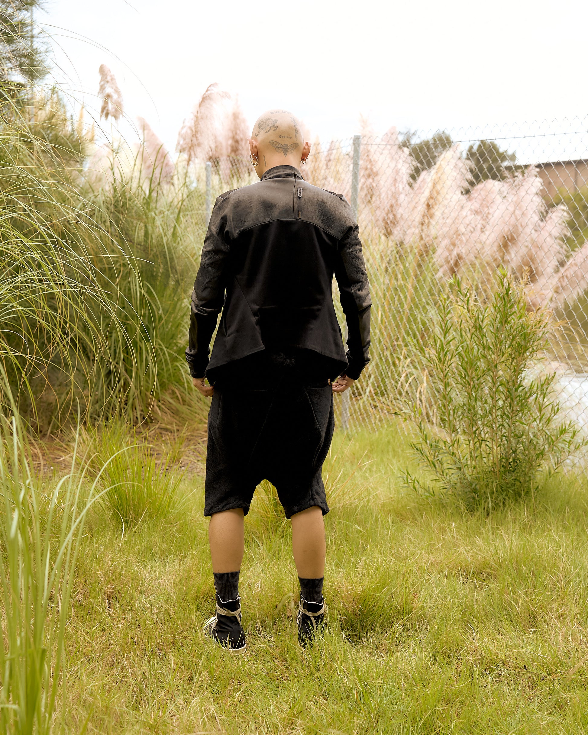 A person with a shaved head faces away, wearing a black Boris Bidjan Saberi jacket and shorts in a grassy area.