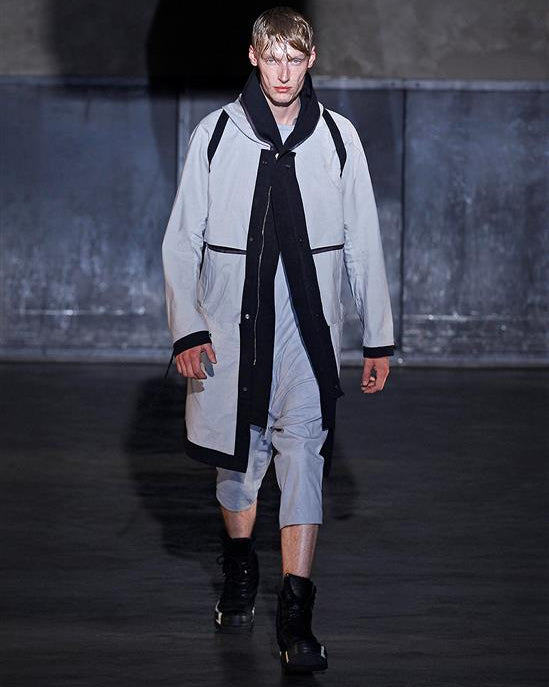 Model wearing a stylish Boris Bidjan Saberi outfit with a long coat and black accents on a dark runway.