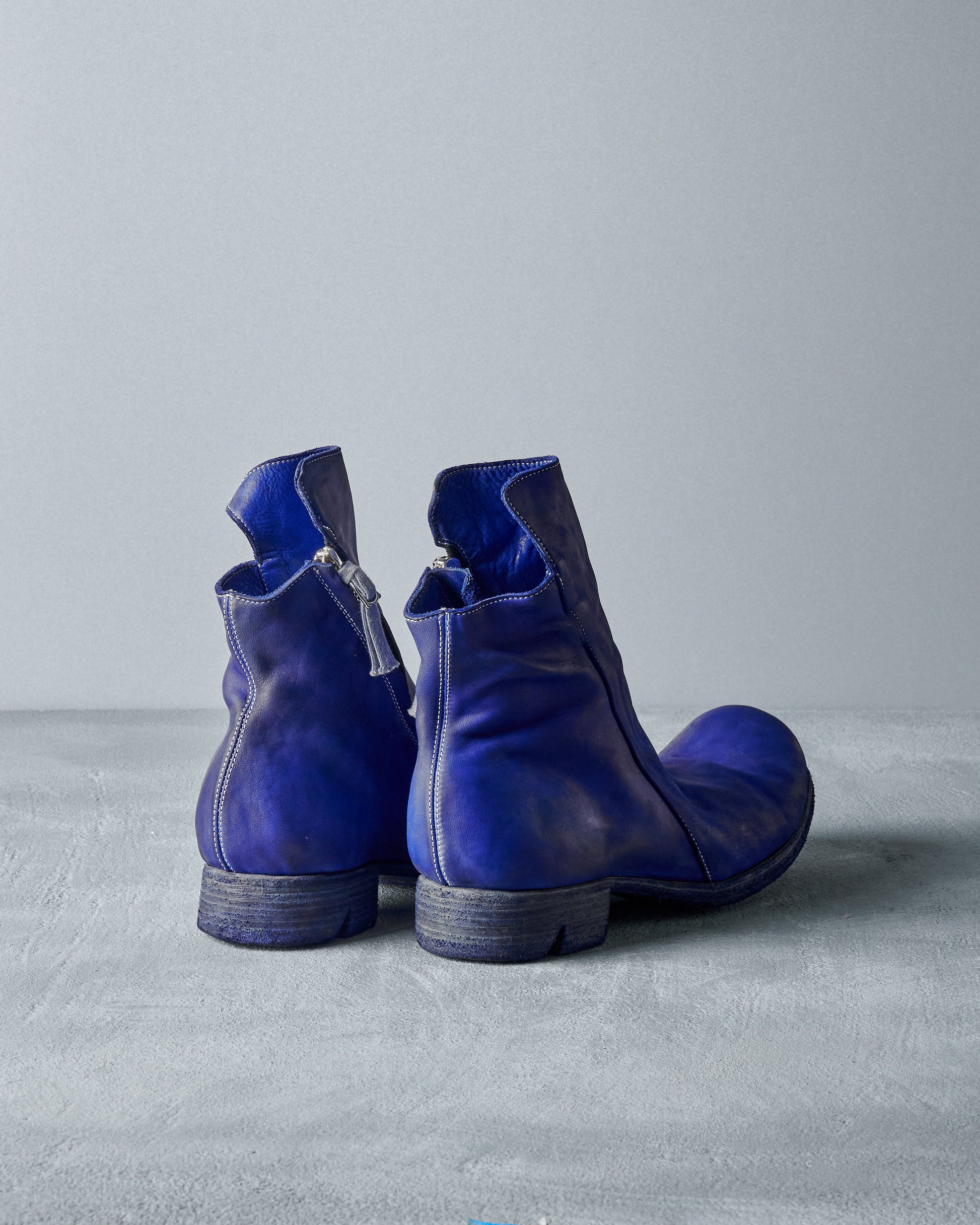 Pair of vibrant blue leather Boris Bidjan Saberi ankle boots with a slightly distressed finish, positioned on a smooth, light grey surface.