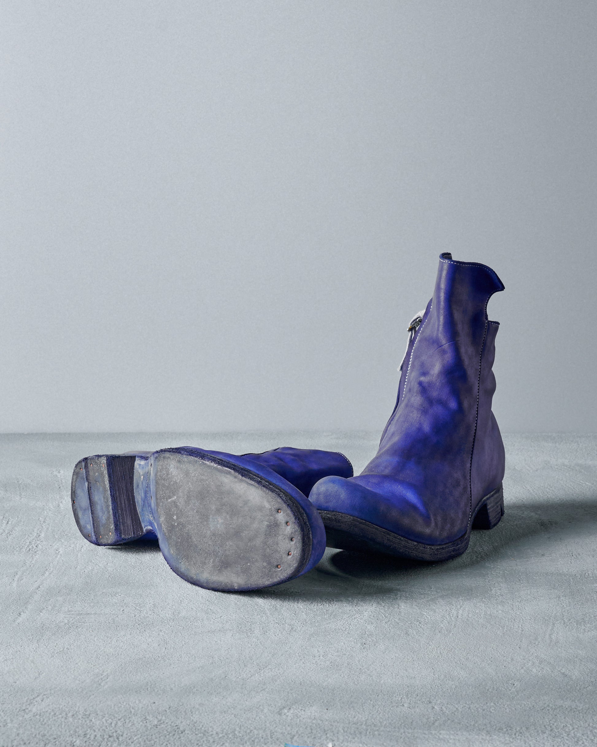 A pair of blue leather Boris Bidjan Saberi boots on a grey surface; one boot stands upright, and the other lies on its side.