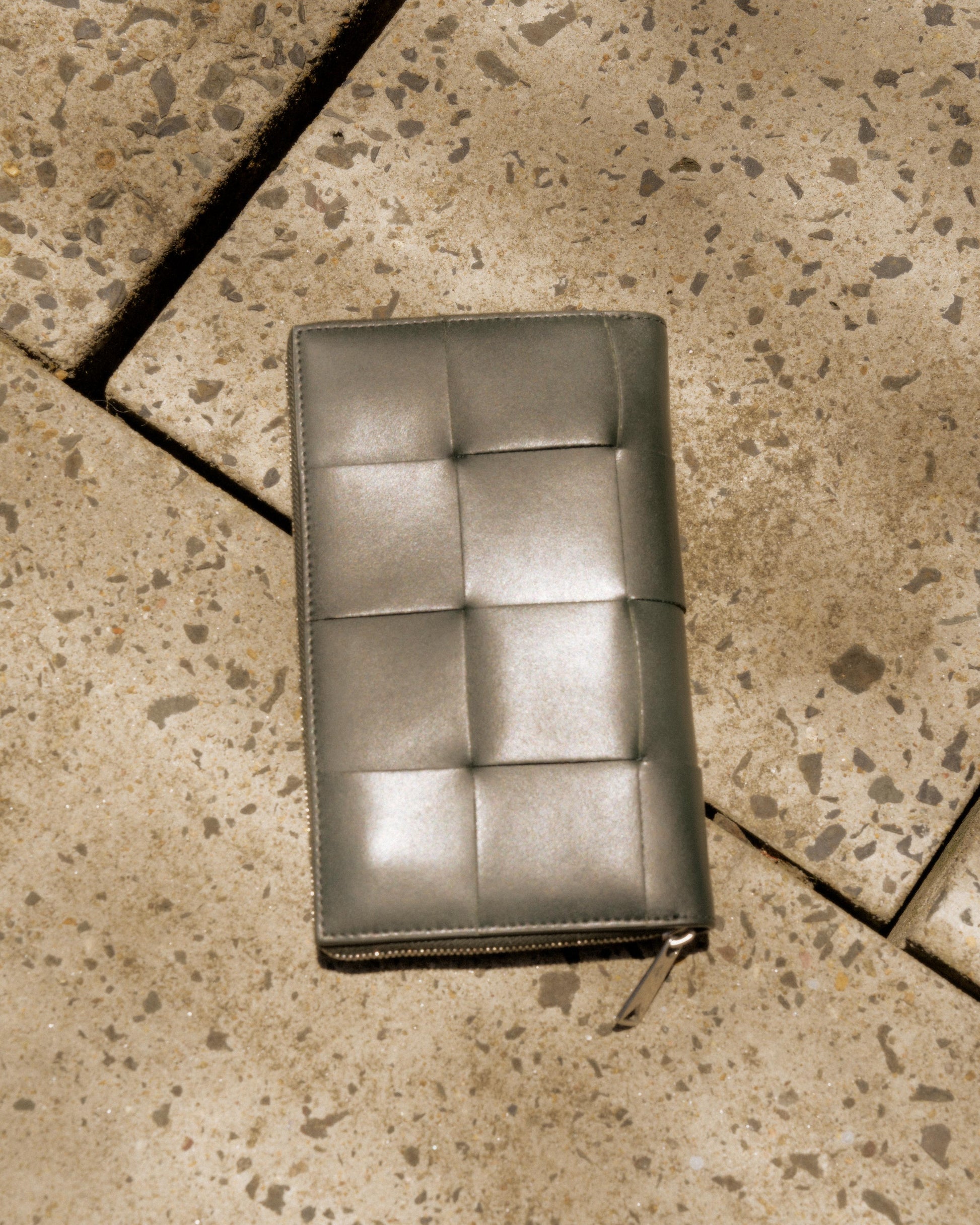 A grey Bottega Veneta intrecciato-woven leather bifold wallet with a side zipper, lying on a textured surface.