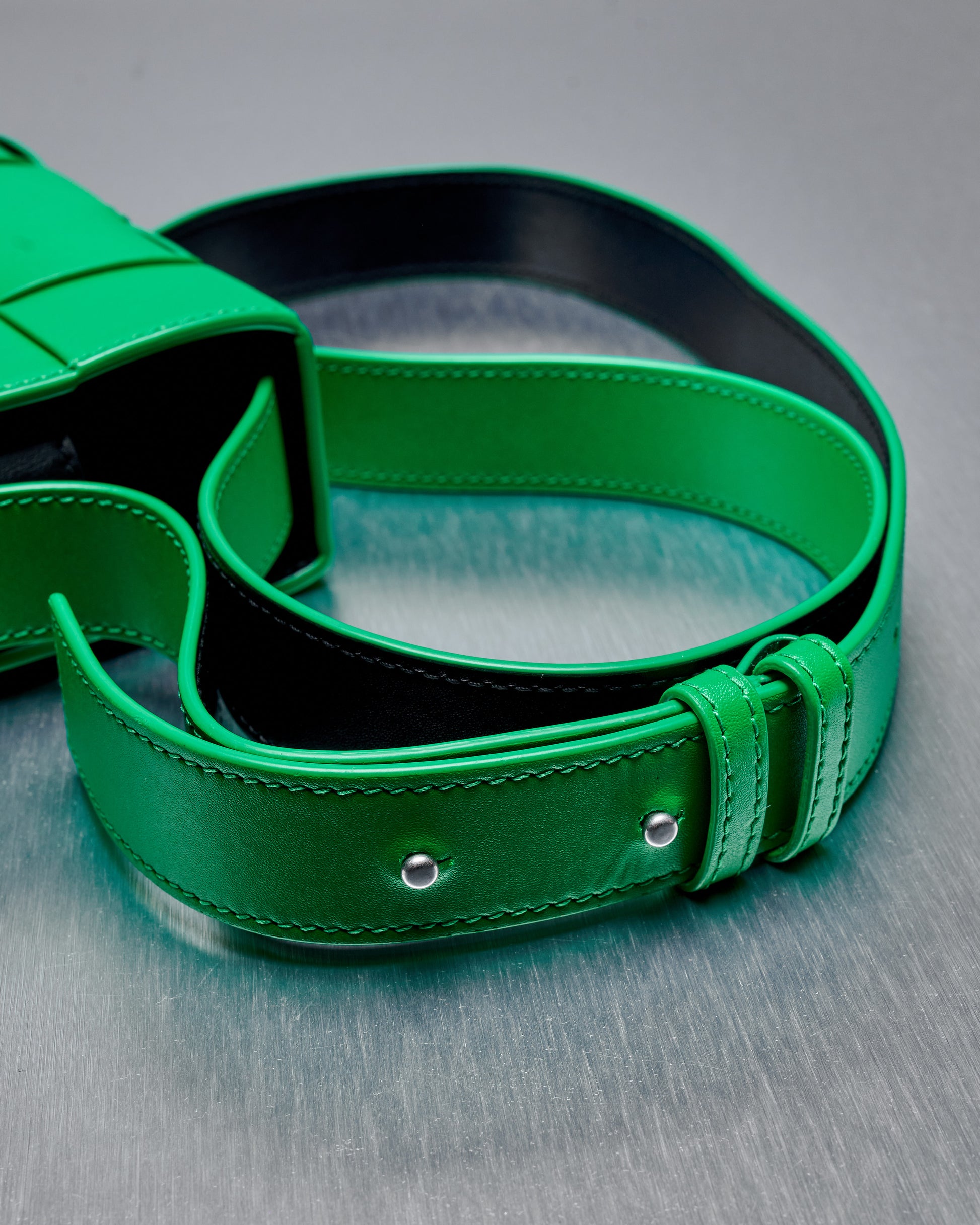 A vibrant green leather Bottega Veneta bag strap with double holes and silver rivets is coiled on a textured grey surface.