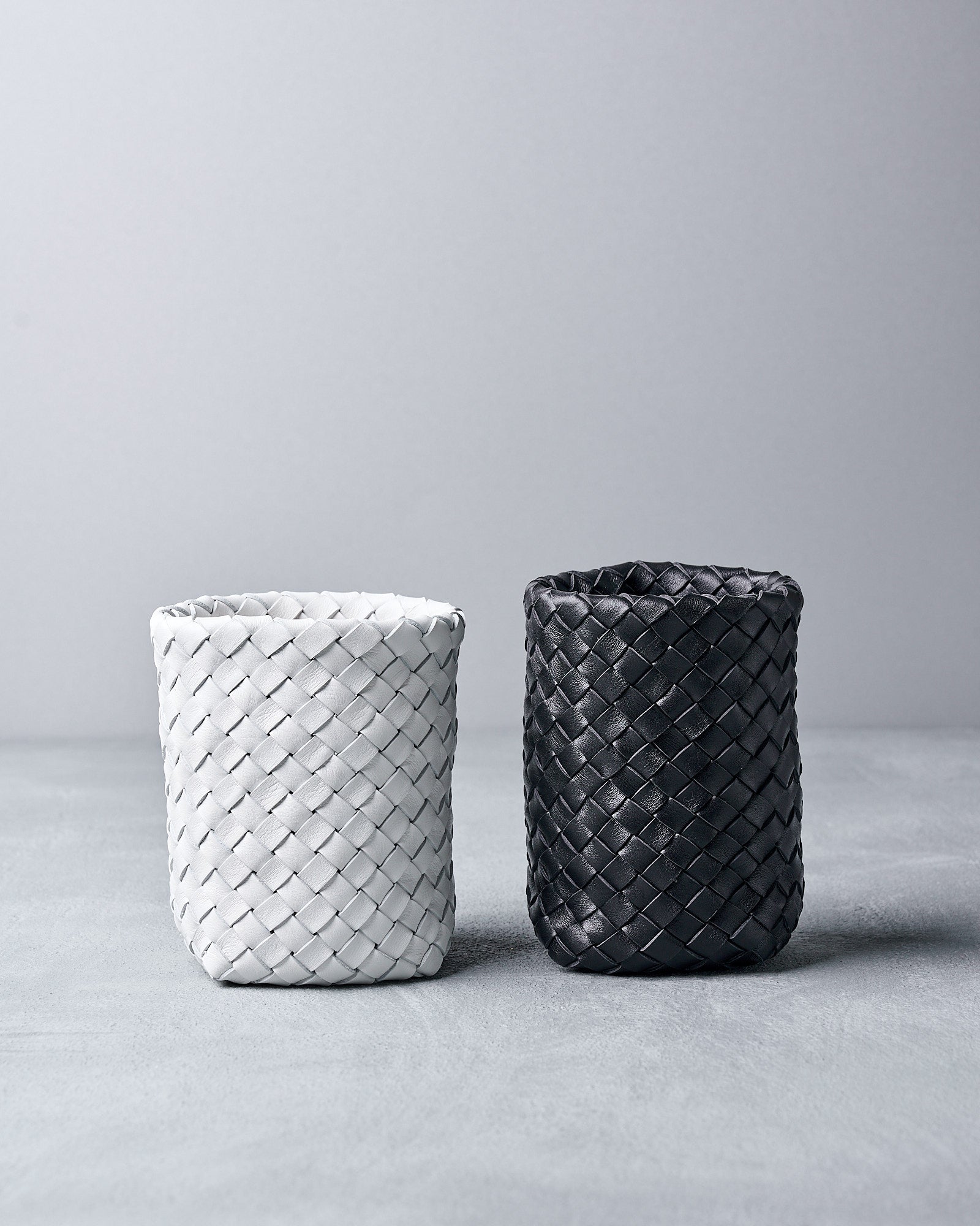 Two woven leather containers for a board game from Bottega Veneta, one white and one black, on a gray surface with a light gray background.
