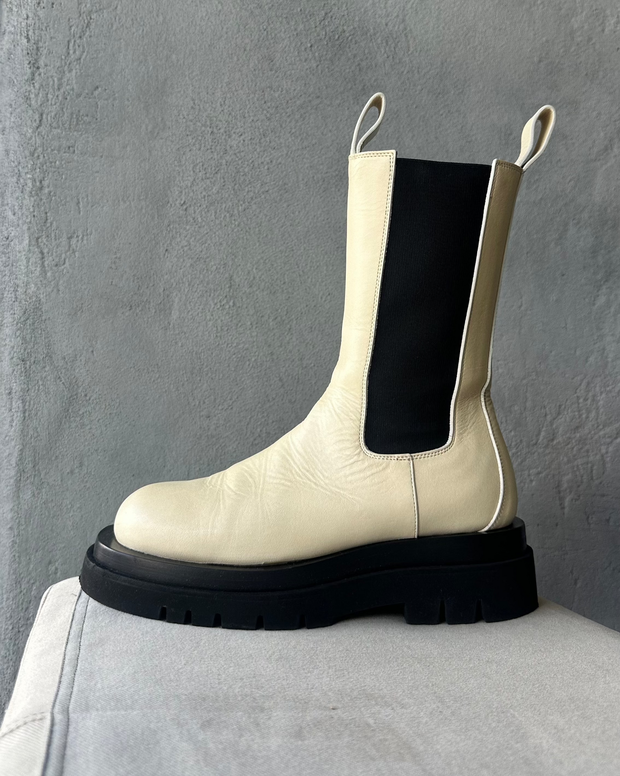 White Bottega Veneta Chelsea boot with black sole on a gray surface against a gray background
