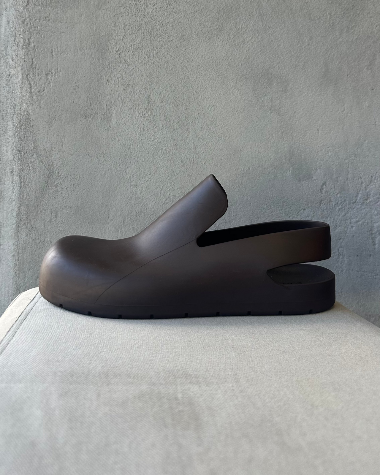 Brown Bottega Veneta clog shoe on a light gray cushion against a textured gray wall