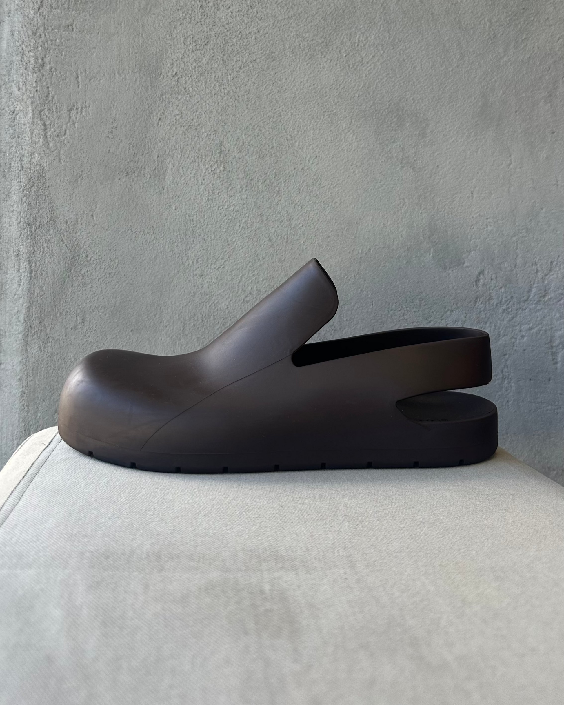 Brown Bottega Veneta clog shoe on a light gray cushion against a textured gray wall
