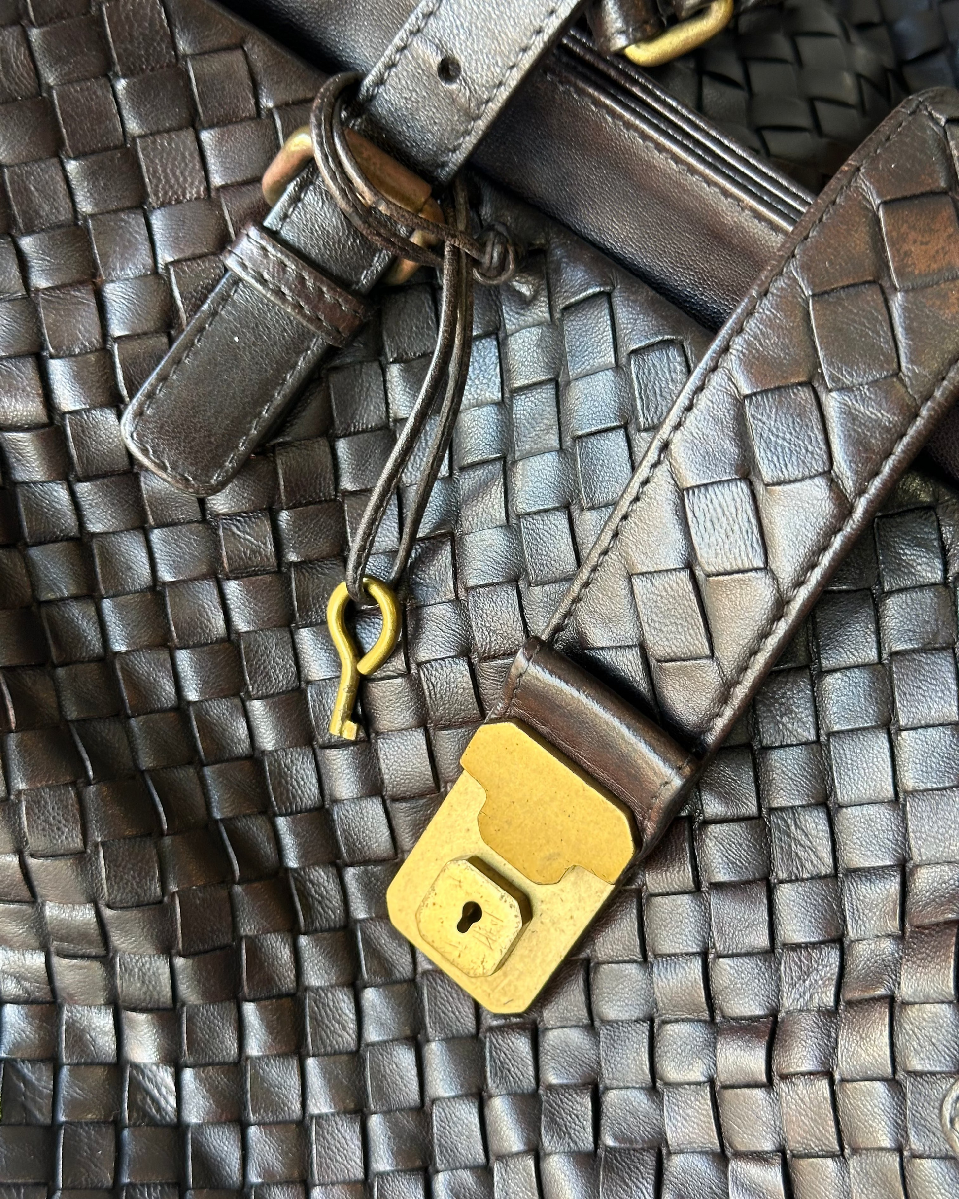 Close-up of a woven leather Bottega Veneta bag with a gold padlock and keychain on a textured surface.