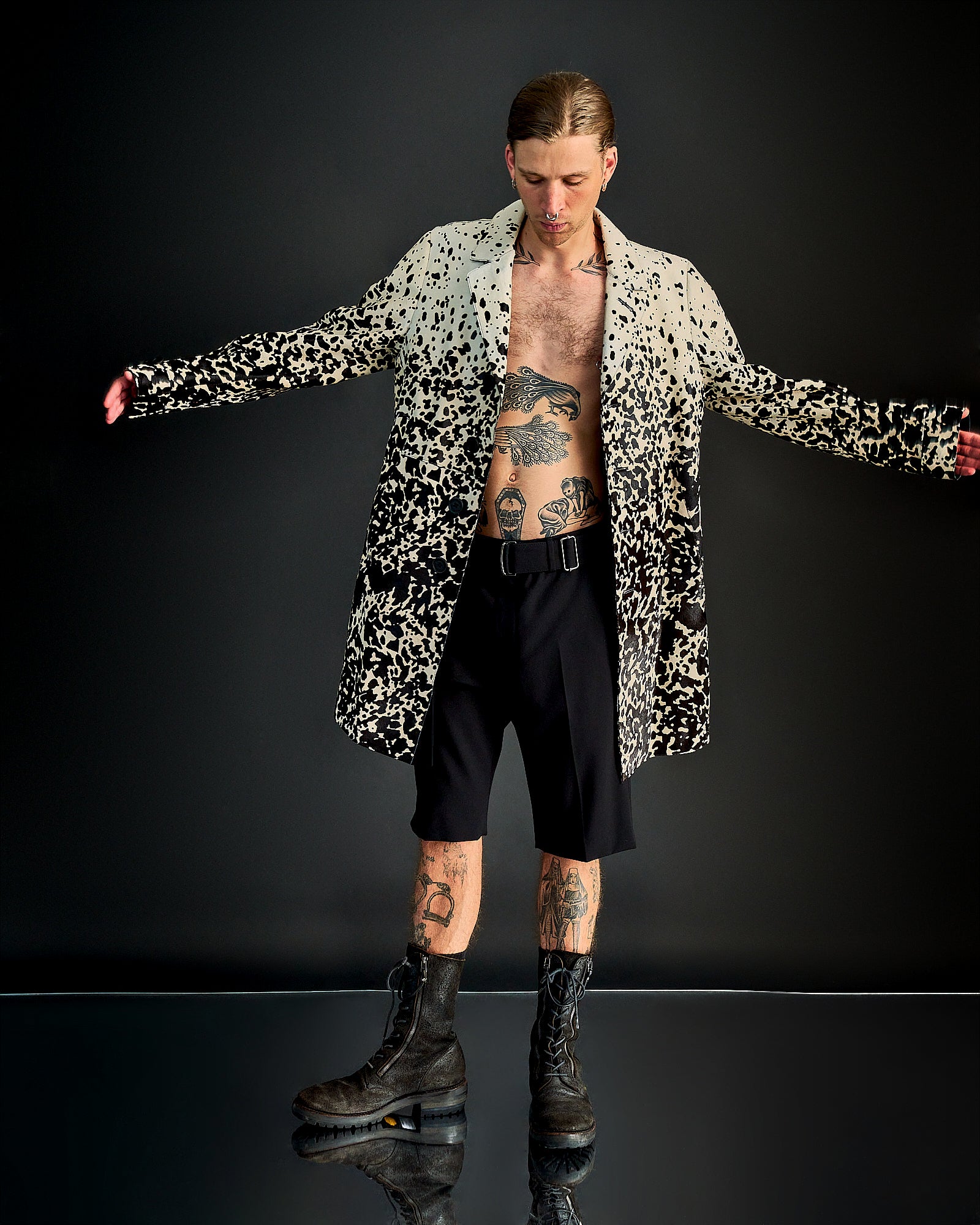 A person poses with arms outstretched, wearing a black and white speckled Burberry coat, black shorts, and boots. 