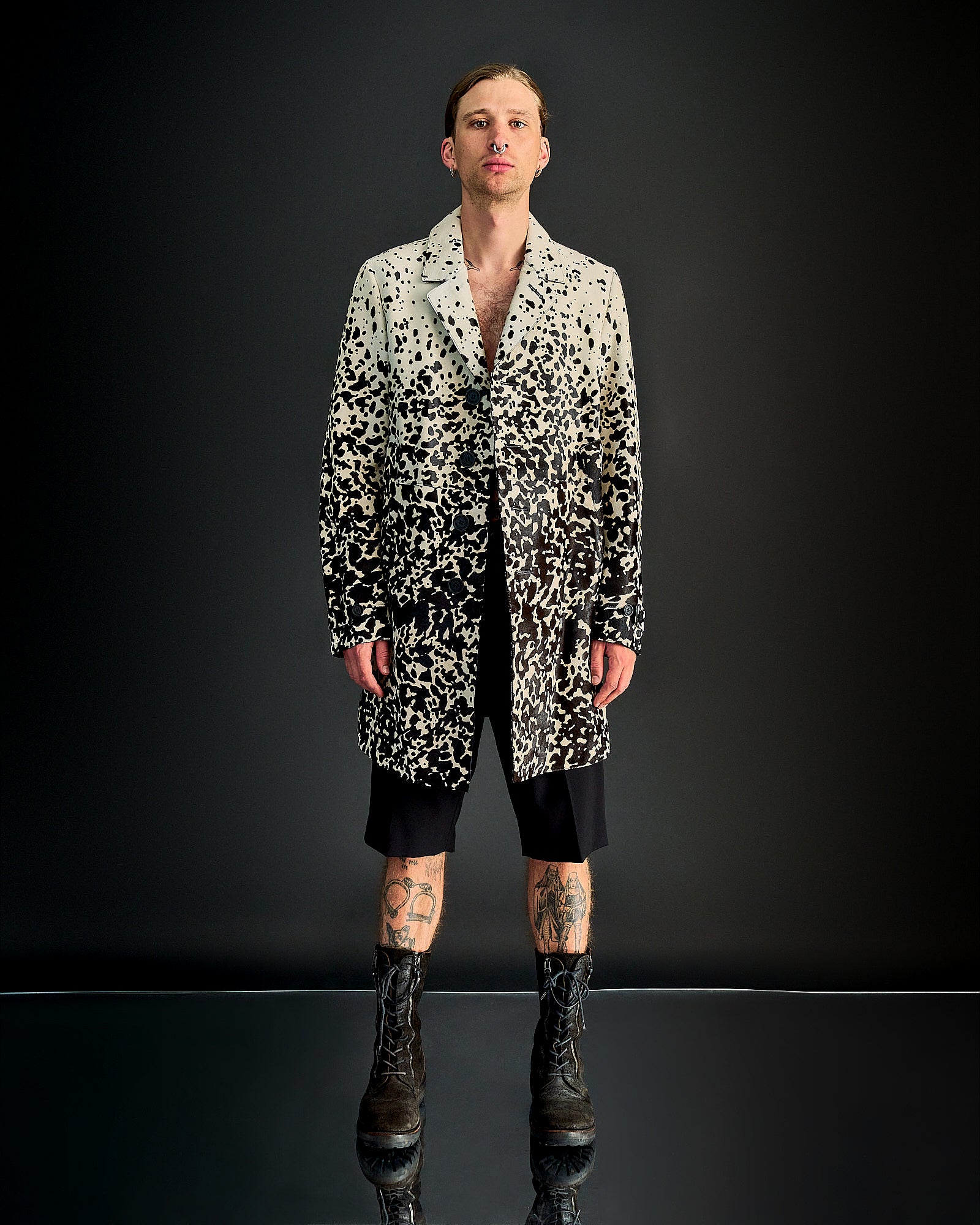 A person stands against a dark background wearing a black and white speckled Burberry coat, black shorts, and rugged boots. Tattoos are visible on their legs