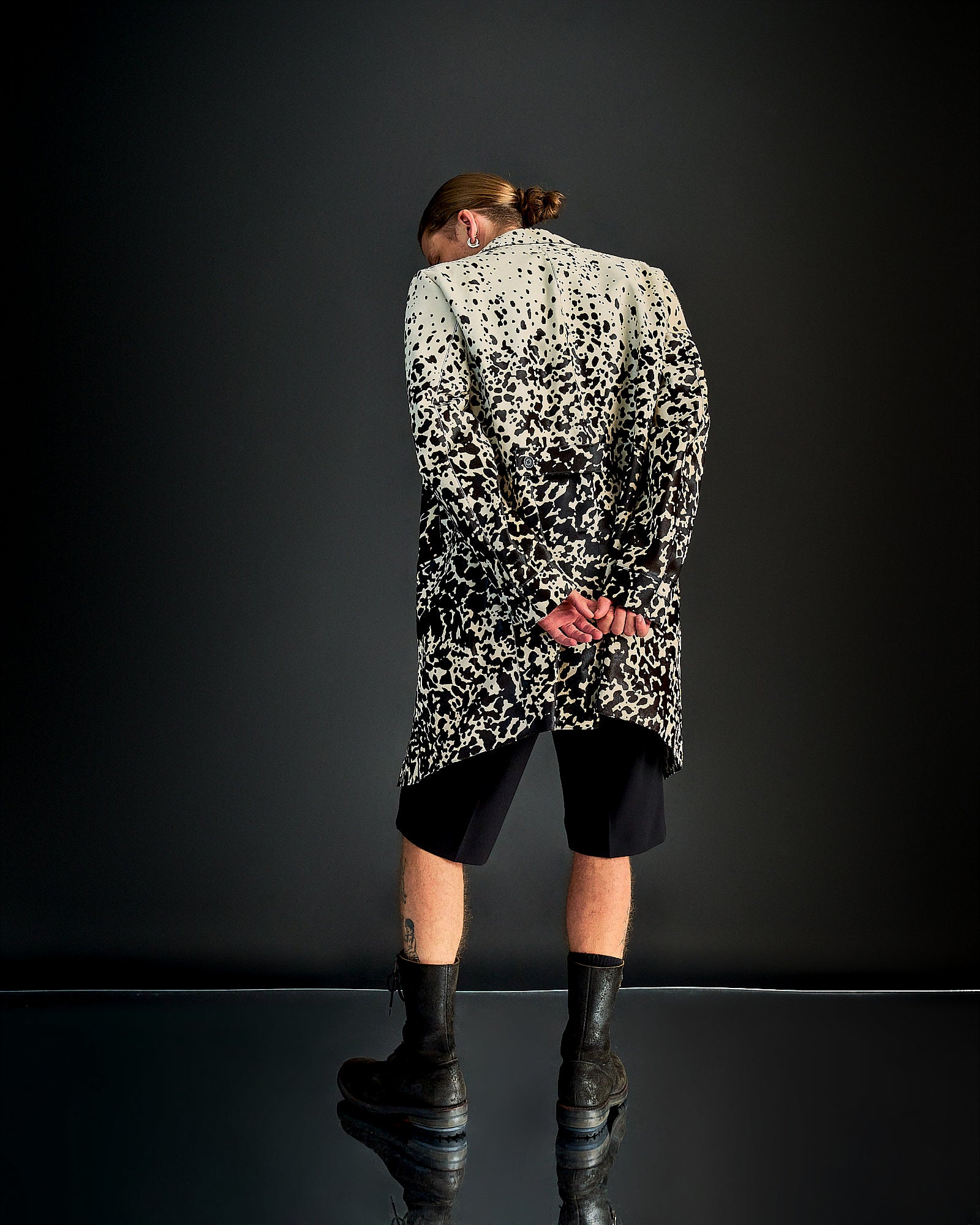 A person with a bun stands back to the camera, hands clasped, wearing a black and white speckled Burberry coat, black shorts, and boots against a dark background.