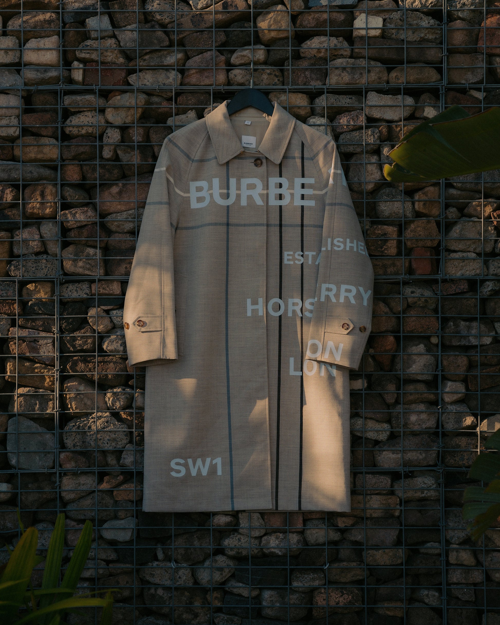 Beige Burberry trench coat with bold white lettering hangs on a metal grid in front of a stone wall, casting soft shadows. Text includes "BURB" and "HORSEFERRY."