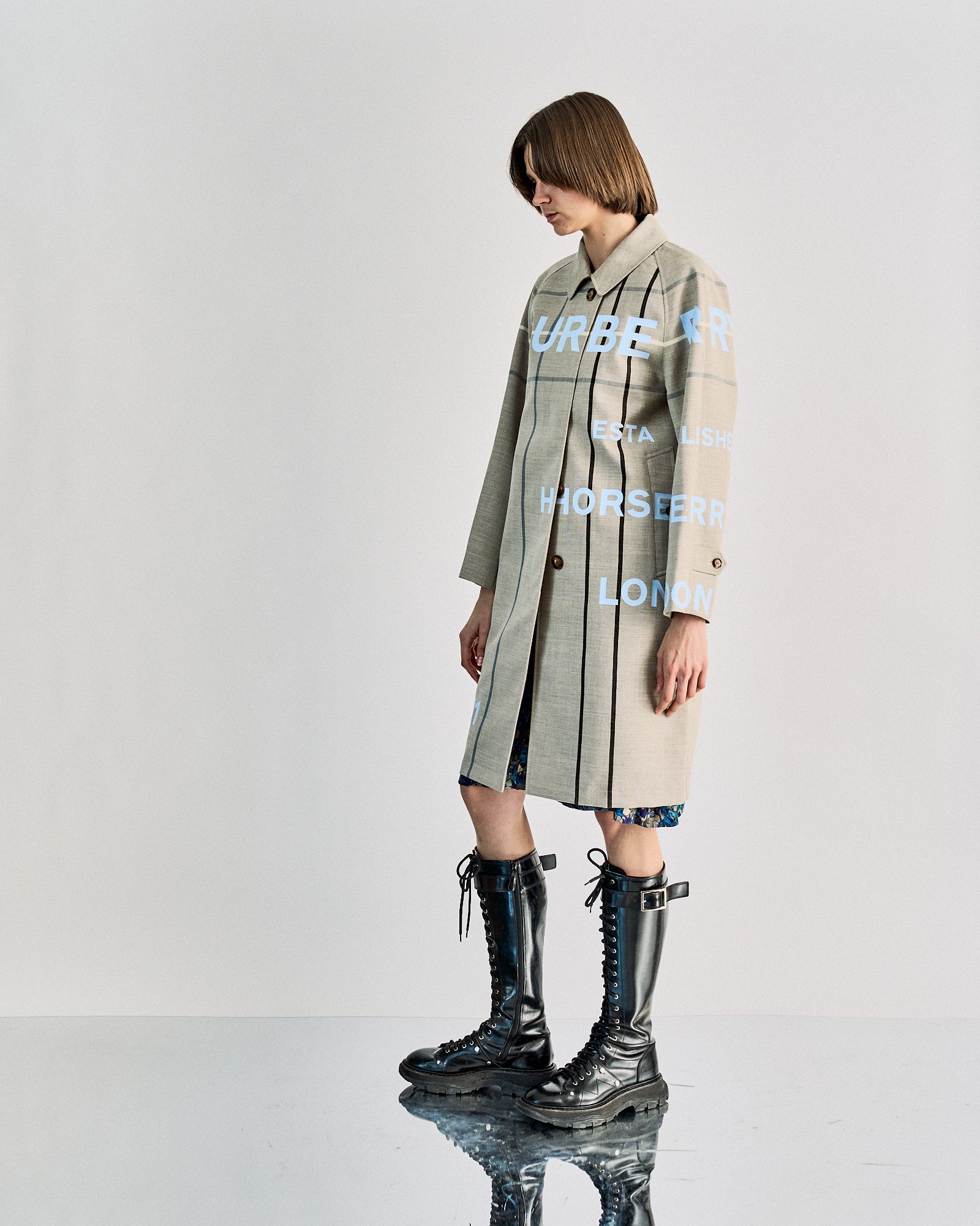 Person with short brown hair wearing a beige Burberry coat with text, standing confidently on a reflective surface. They sport a floral dress and high black boots.