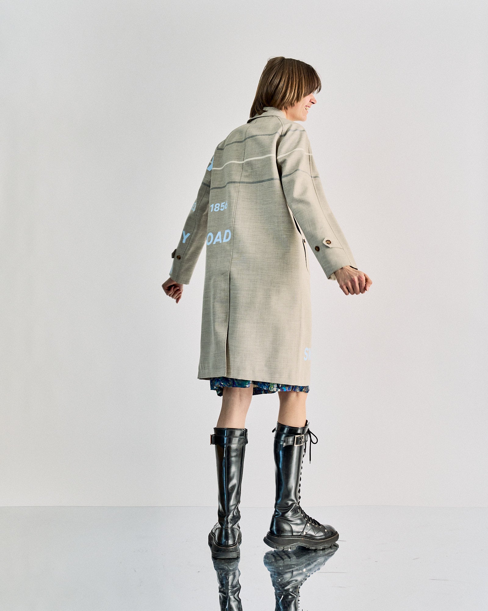 A person stands facing away, wearing a long beige coat with text, over a patterned dress. They have chin-length hair and wear tall, lace-up black boots.