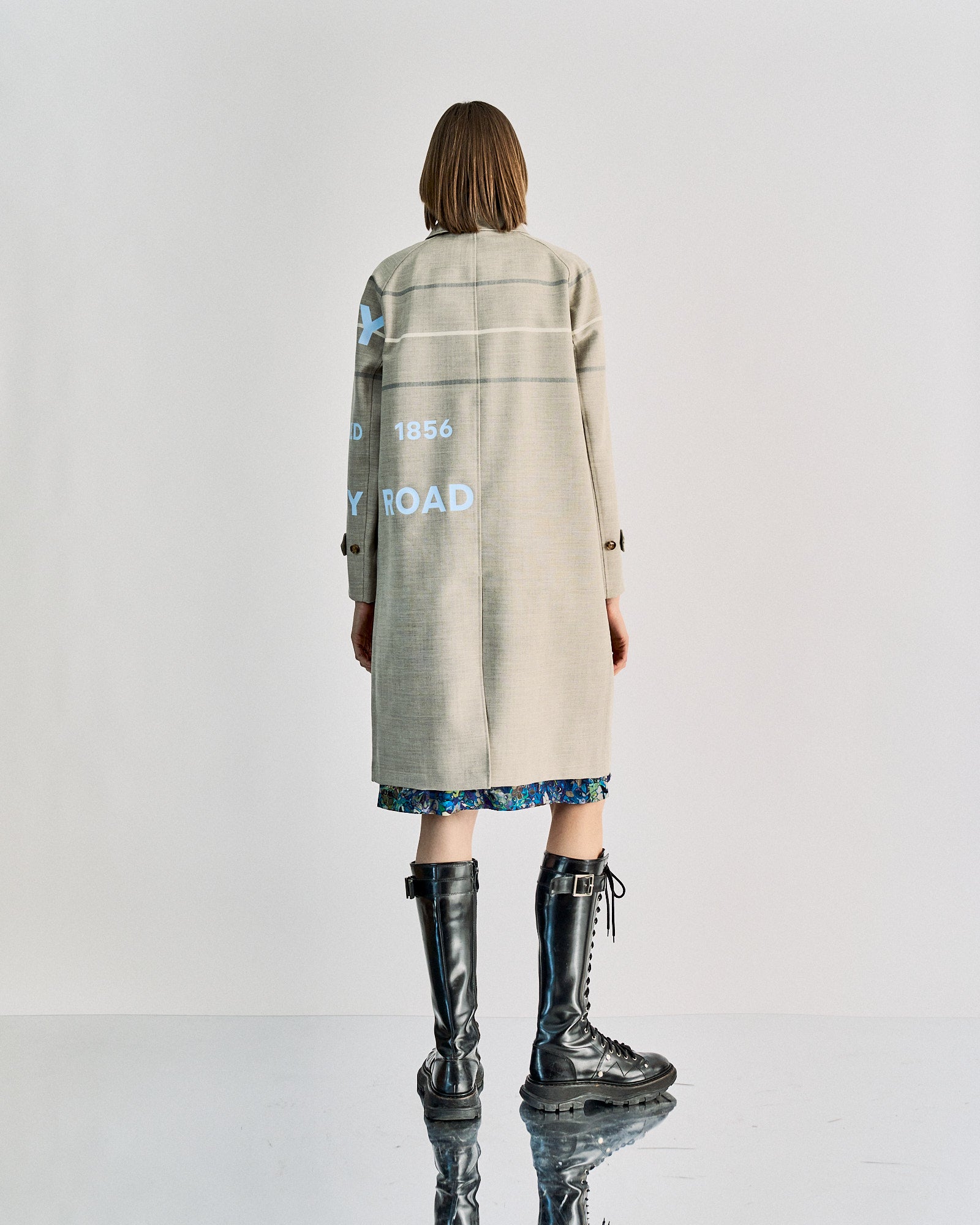 A person stands facing away, wearing a long beige Burberry coat with text, over a patterned dress. They have chin-length hair and wear tall, lace-up black boots.