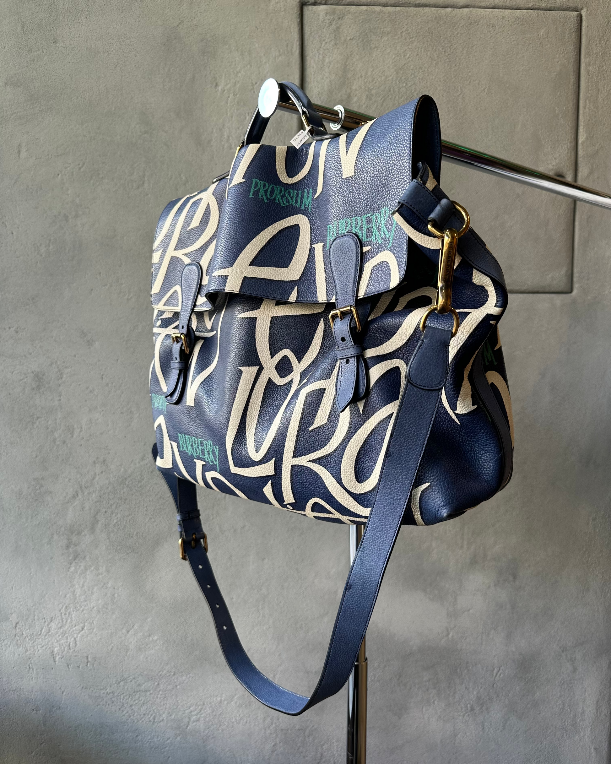 Blue and white patterned Burberry backpack hanging on a wall hook against a gray concrete background