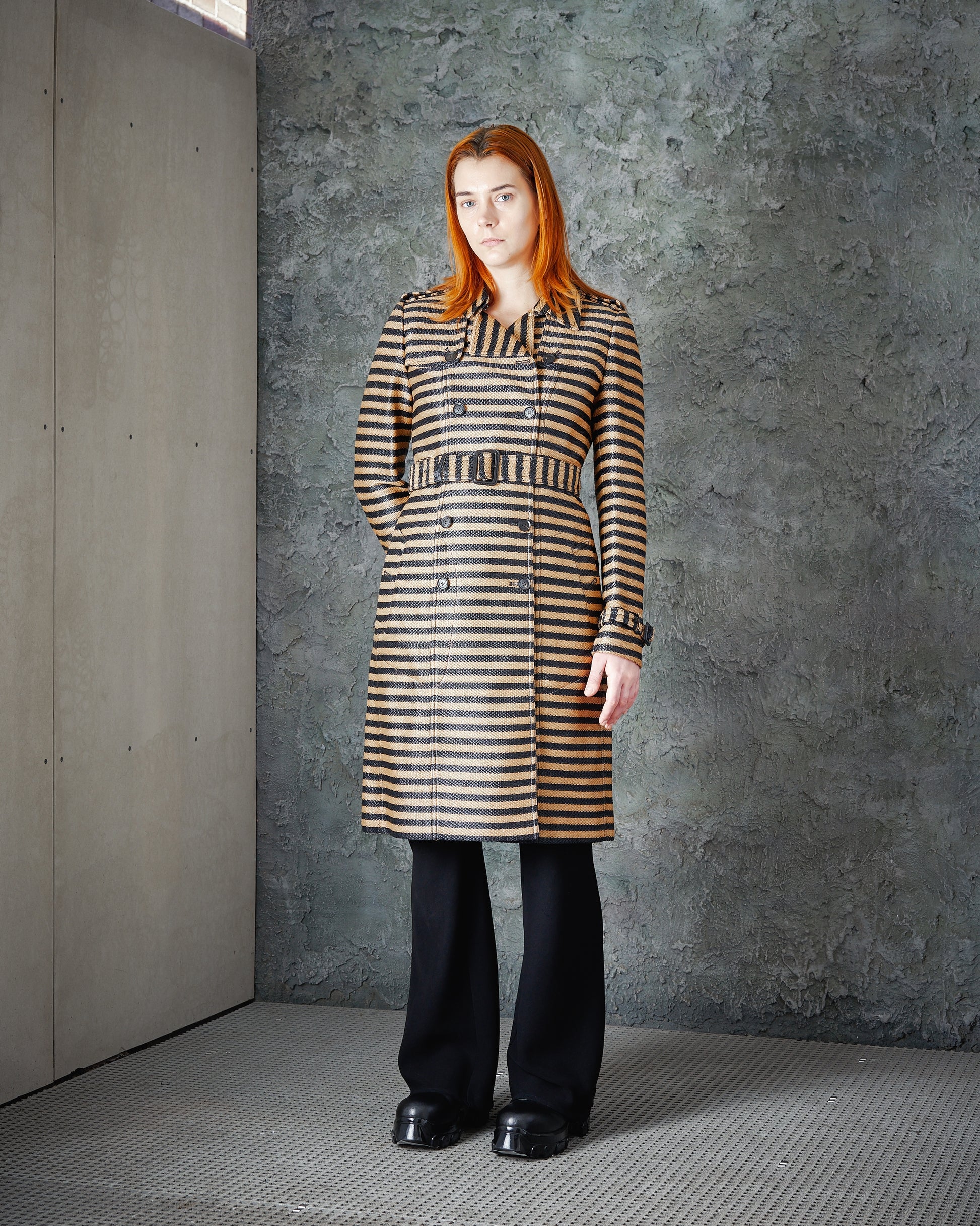 Woman with long red hair in a striped Burberry coat and black pants stands against a textured grey wall.