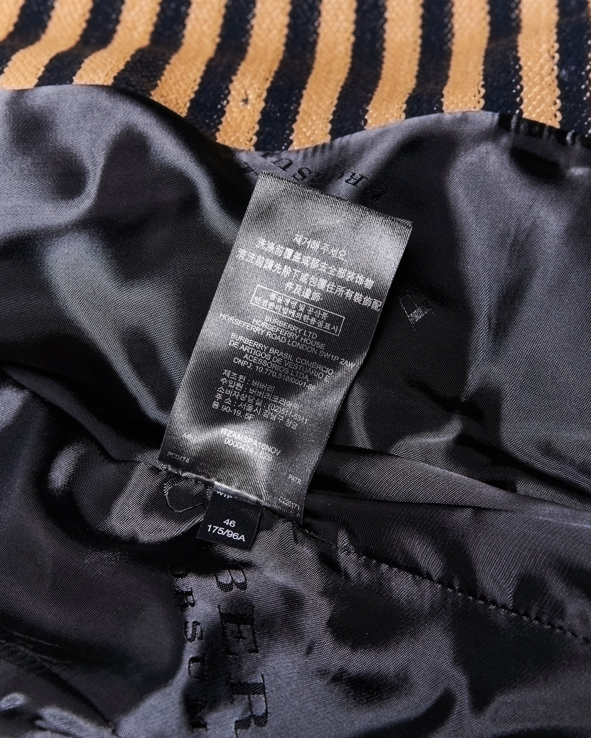 A black Burberry garment label with text in multiple languages including Chinese, English, and others, is attached to a silky black fabric. The fabric lies over a background with gold and black stripes.