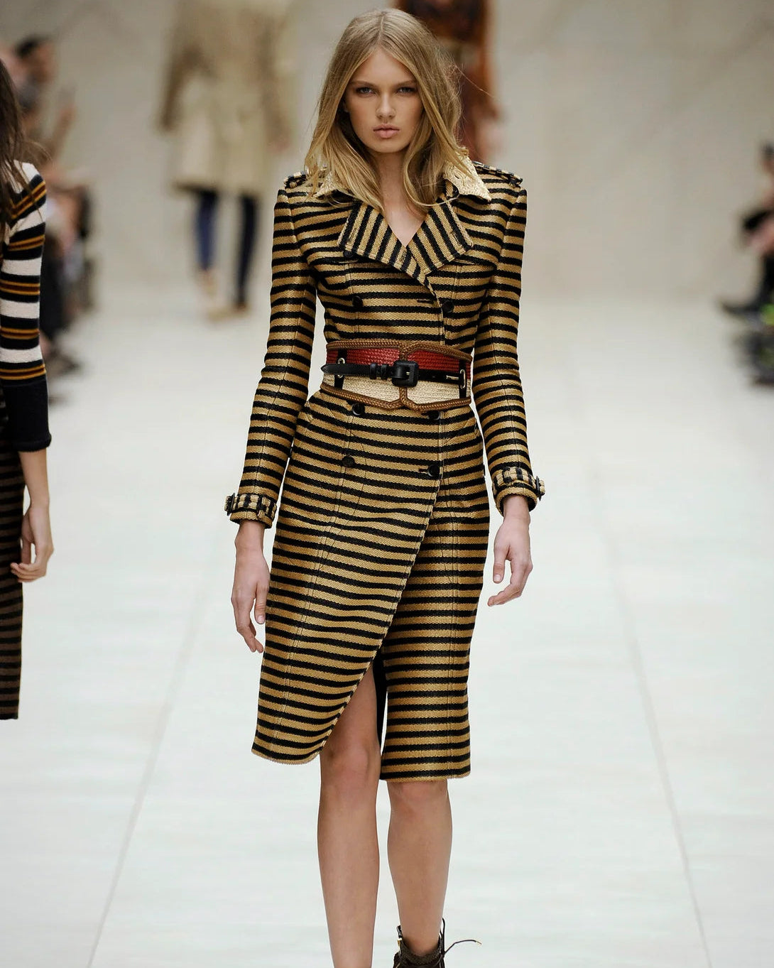 A model walks the runway in a brown and black striped Burberry coat dress, accessorised with a red belt and black boots.