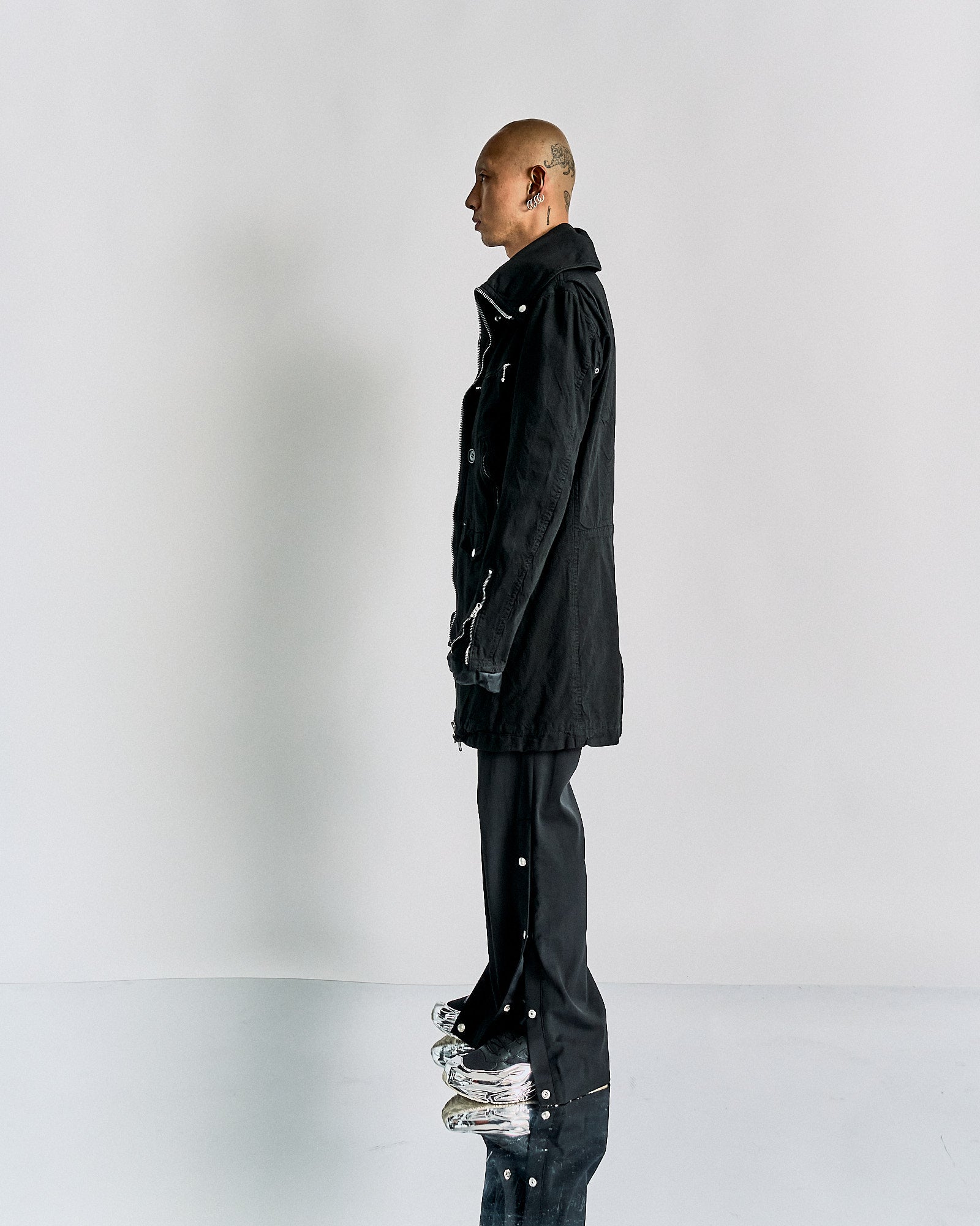 A person stands in profile against a plain background, wearing a long CDG Junya Watanbe black coat and black pants adorned with buttons. Their demeanour is calm and contemplative.