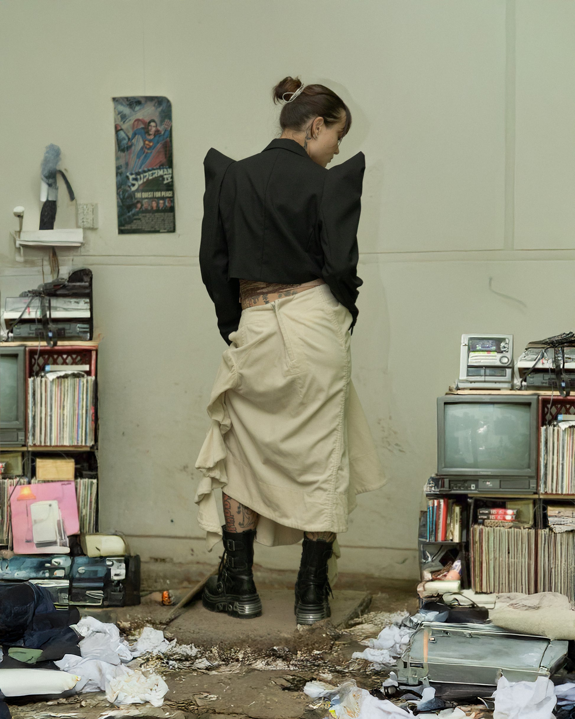 Person in a black Noir Kei Ninomiya cropped blazer and beige skirt standing in a cluttered room with vintage equipment.