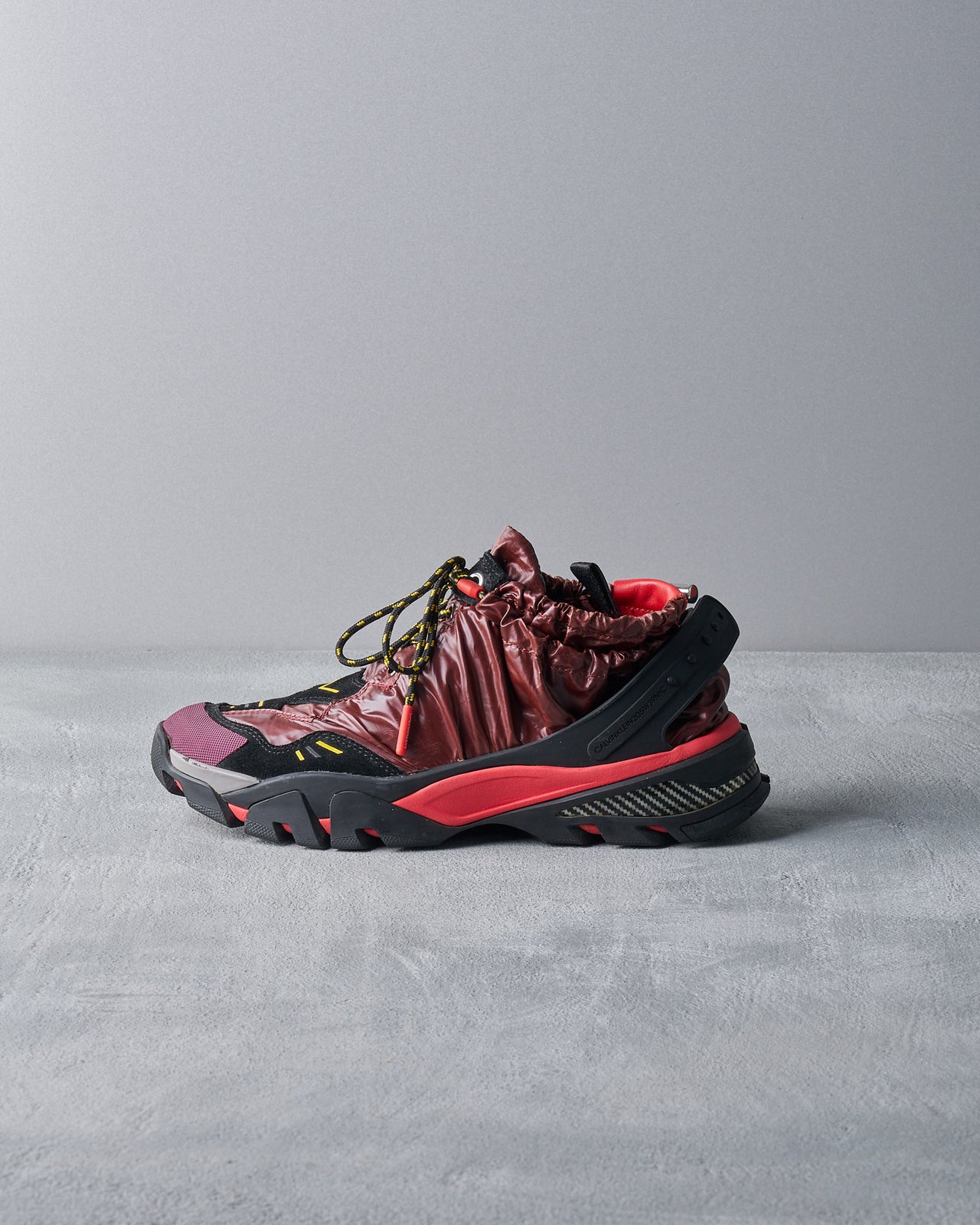 A unique Calvin Klein sneaker combines a glossy maroon upper with black and red accents. It features a rugged sole and drawstring details, casting an edgy, modern vibe.