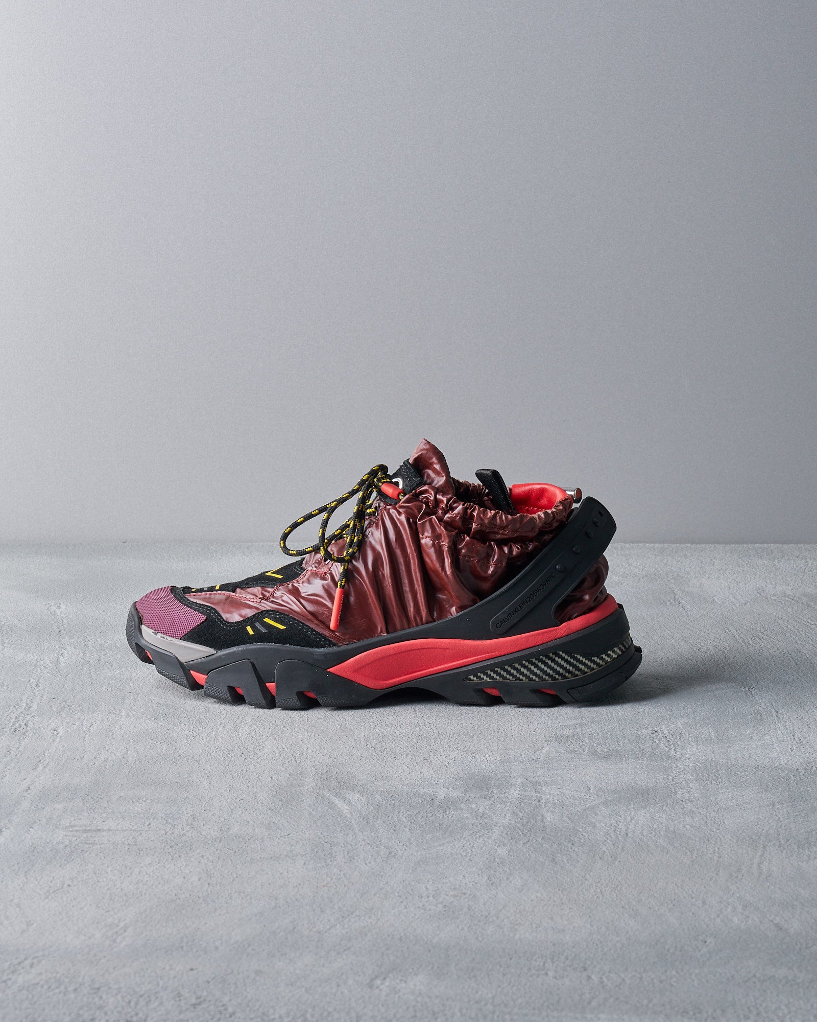 A unique Calvin Klein sneaker combines a glossy maroon upper with black and red accents. It features a rugged sole and drawstring details, casting an edgy, modern vibe.