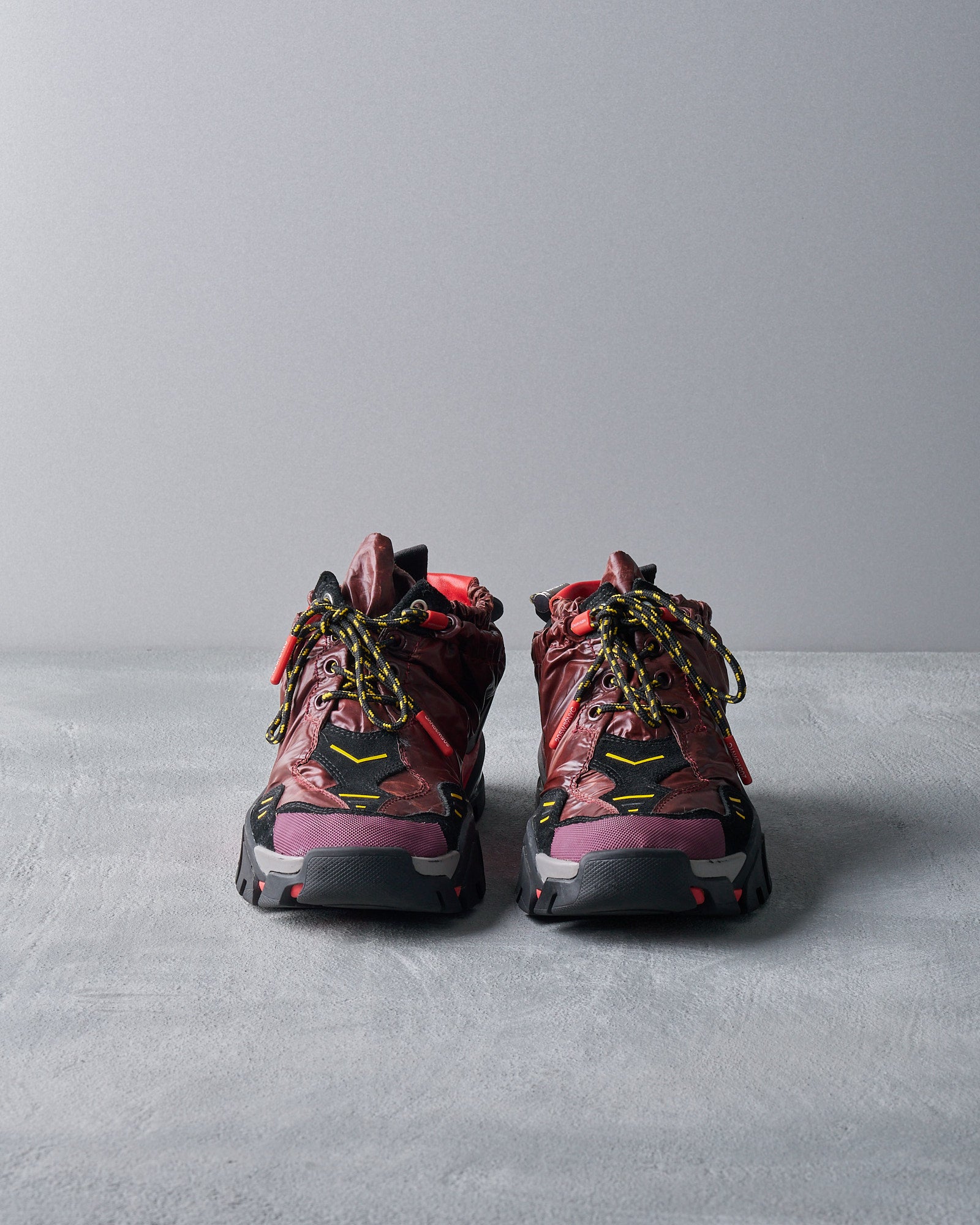 Red and black Calvin Klein hiking boots with yellow laces and accents, placed on a gray textured surface, conveying a rugged and stylish outdoor vibe.