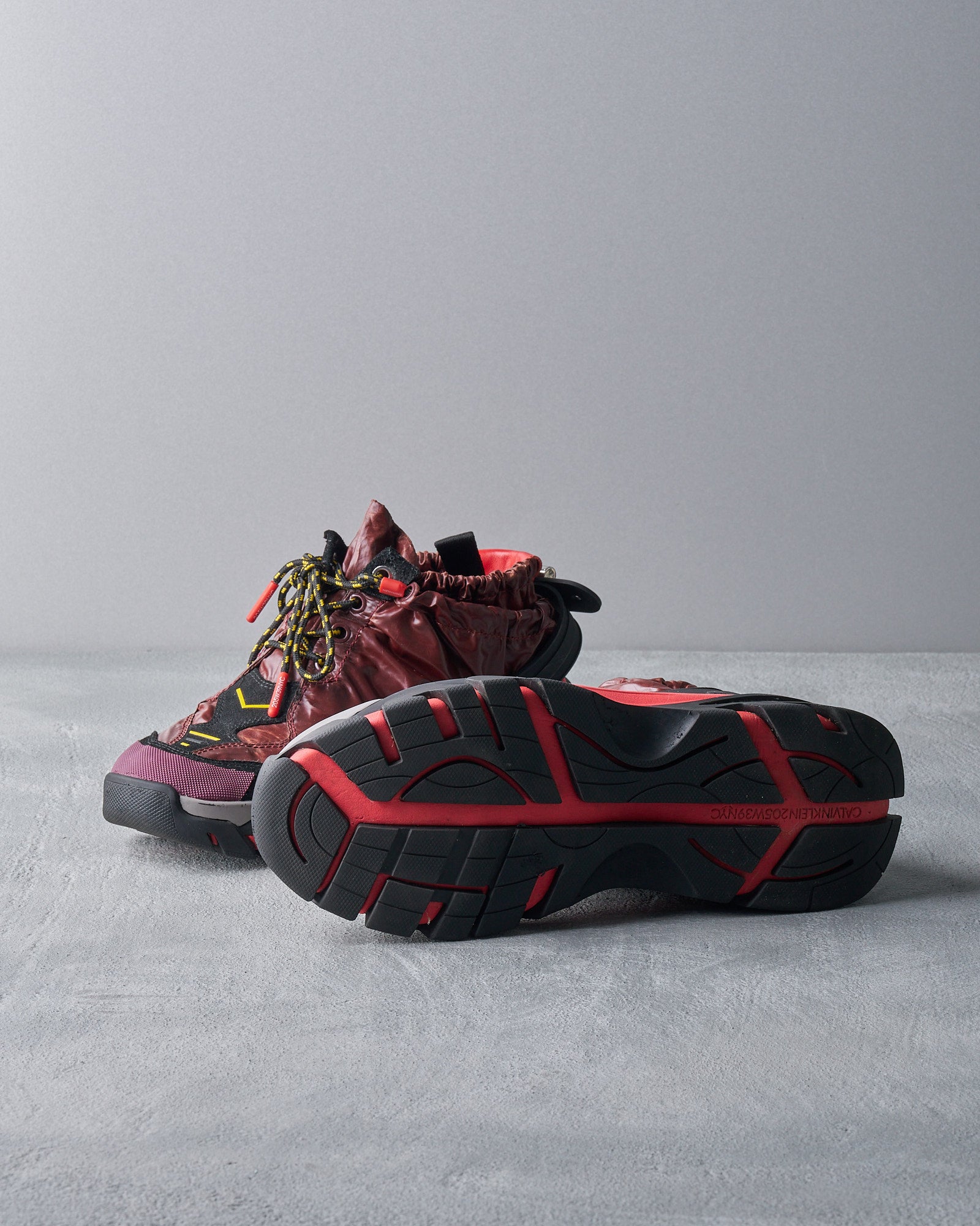 Trendy Calvin Klein sneakers with a rugged design are placed on a light surface. One shoe is upright, showcasing maroon tones and colorful laces; the other shows a bold red and black sole.