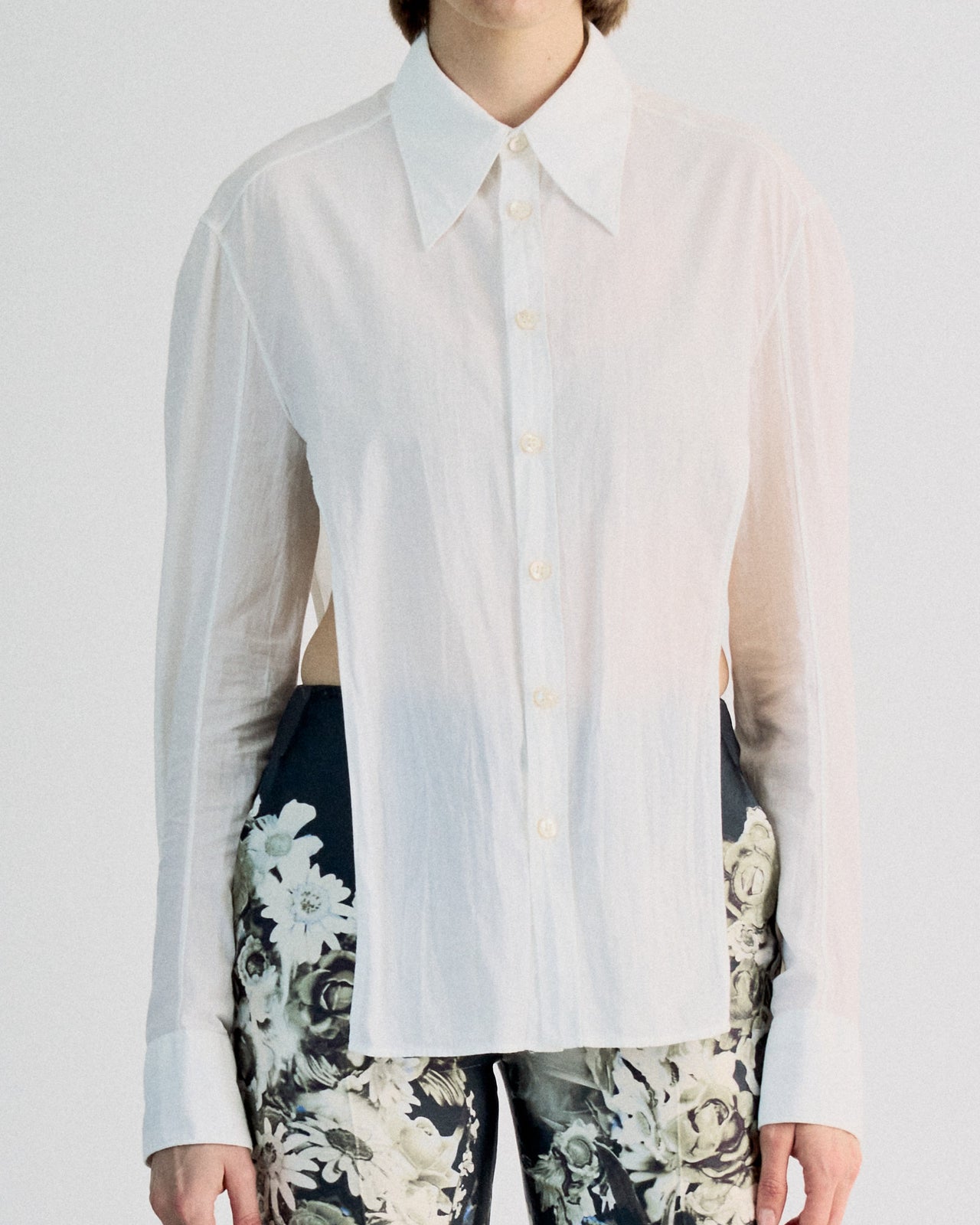 A person wearing a white unbuttoned Carol Christian Poell shirt, floral shorts, and tall black lace-up boots stands on a reflective surface, evoking a modern, edgy vibe.
