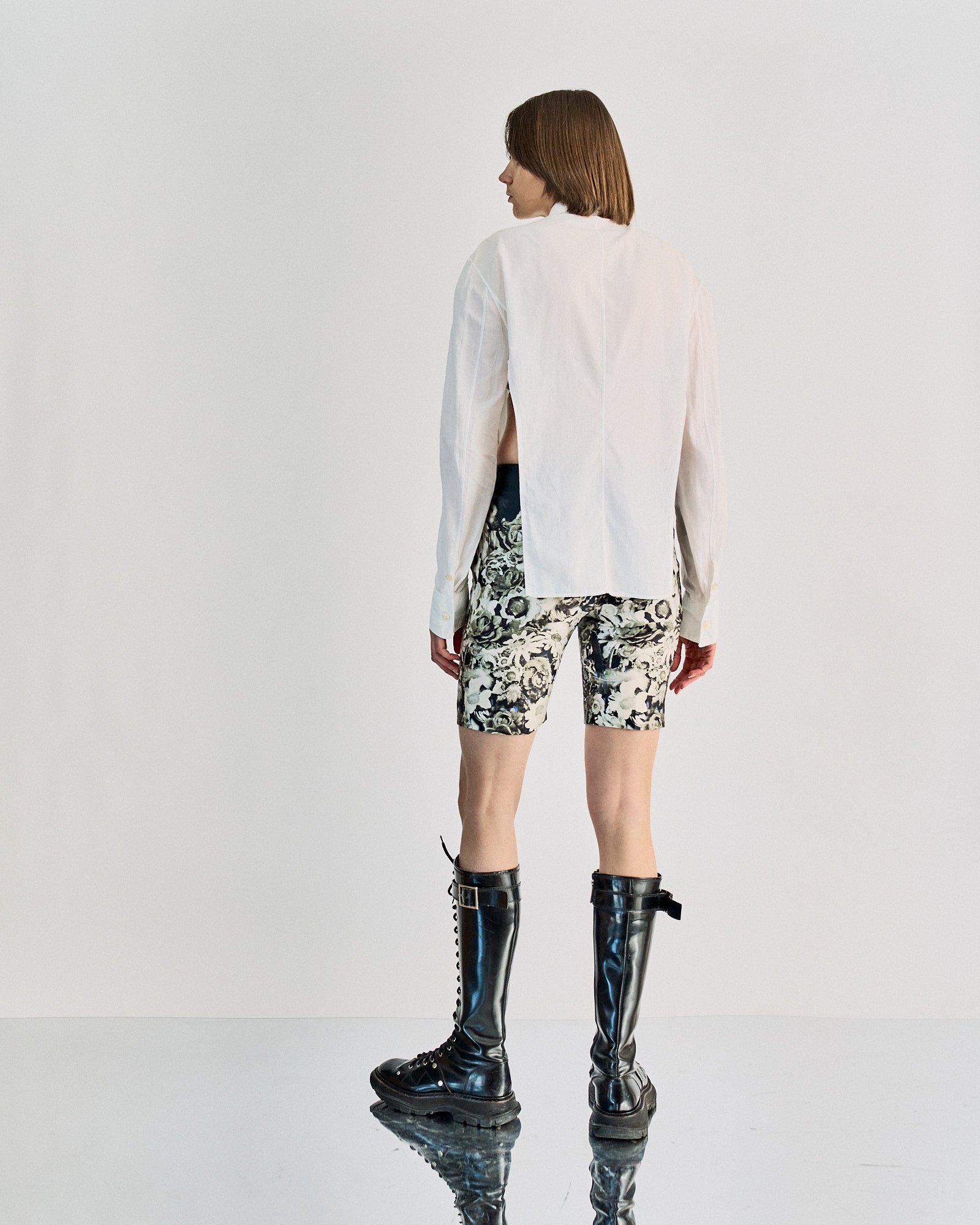A person with shoulder-length hair faces away, wearing a sheer white Carol Christian Poell shirt, floral shorts, and tall black boots. 