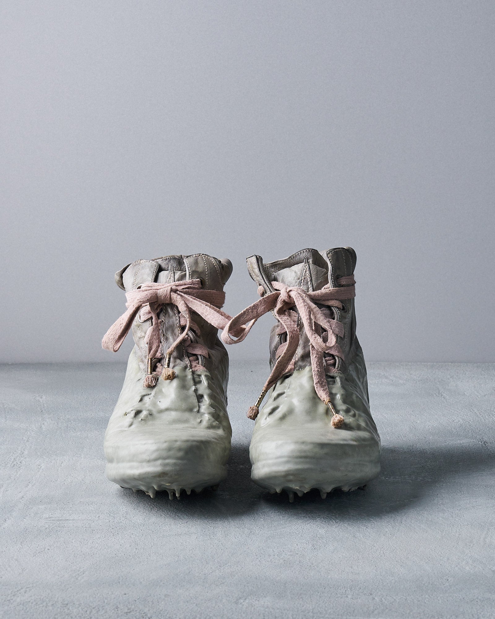 Worn grey Carol Christian Poell boots with pink laces and glossy, rugged soles, resembling a melt or fusion of materials. They rest on a textured grey surface.