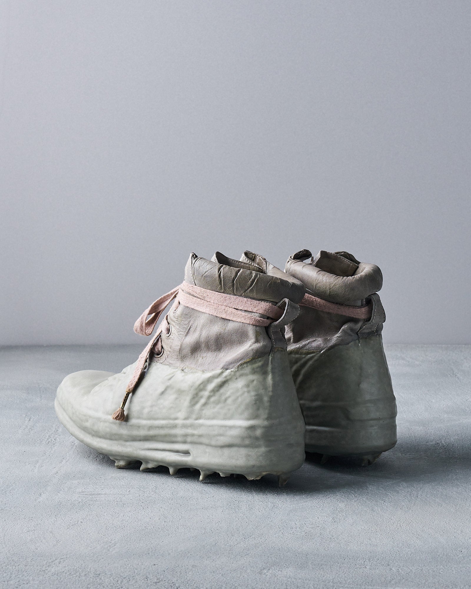 Worn grey Carol Christian Poell boots with pink laces and glossy, rugged soles, resembling a melt or fusion of materials. They rest on a textured grey surface.
