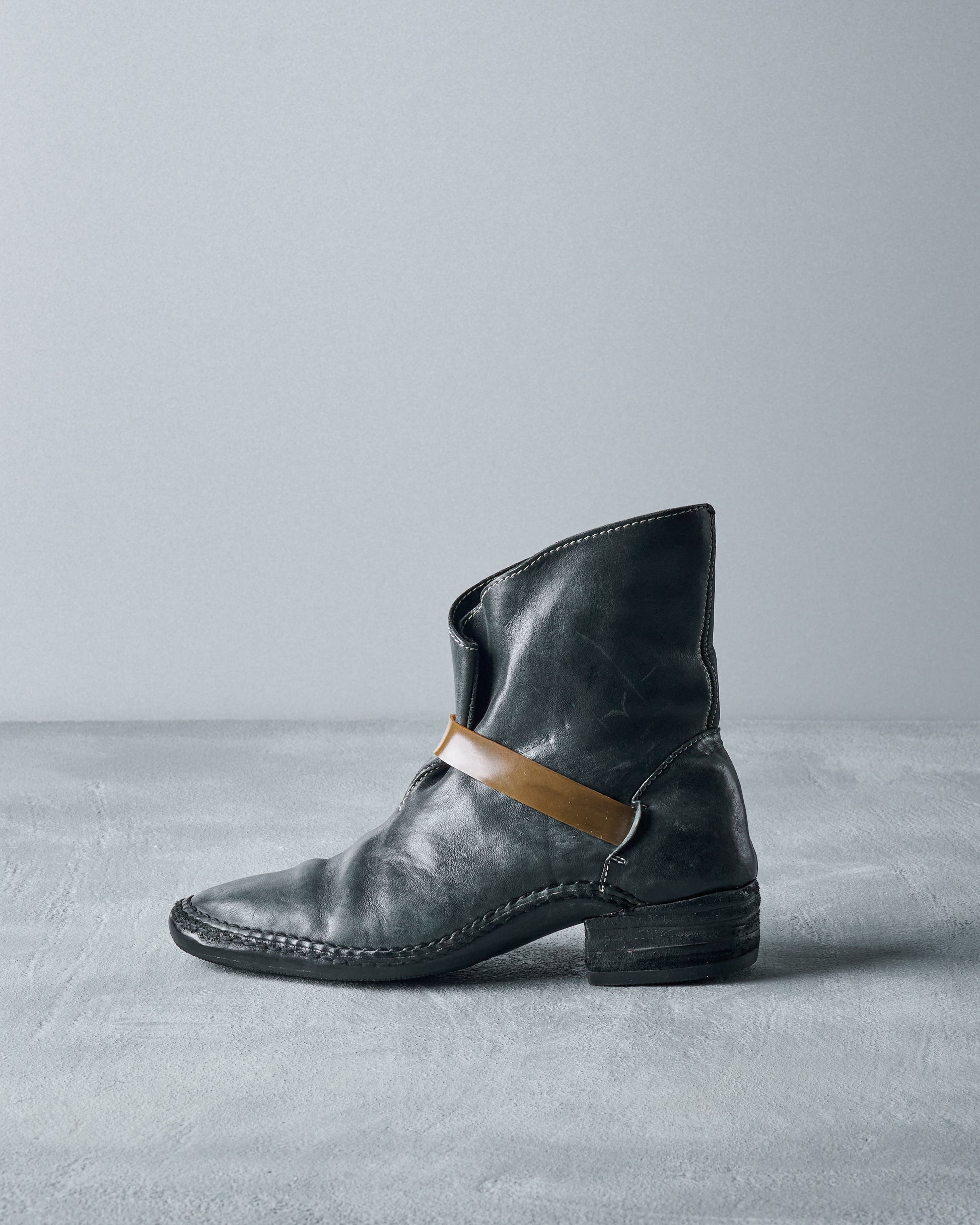 A black leather Carol Christian Poell lace-up boot with a thick rubber band wrapping around the ankle, positioned against a grey background.