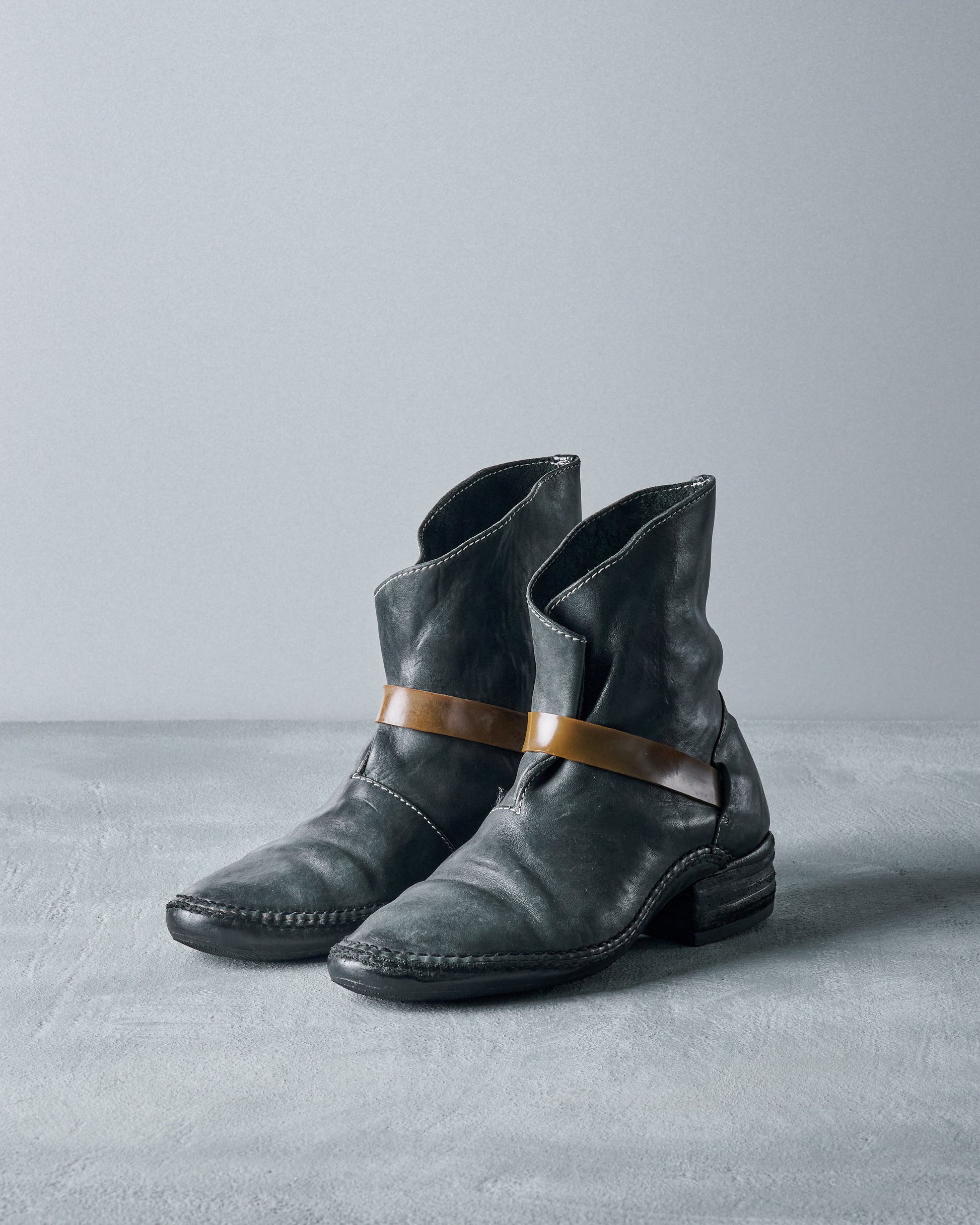 Black leather Carol Christian Poell lace-up boots with a thick rubber band wrapping around the ankle, positioned against a grey background.